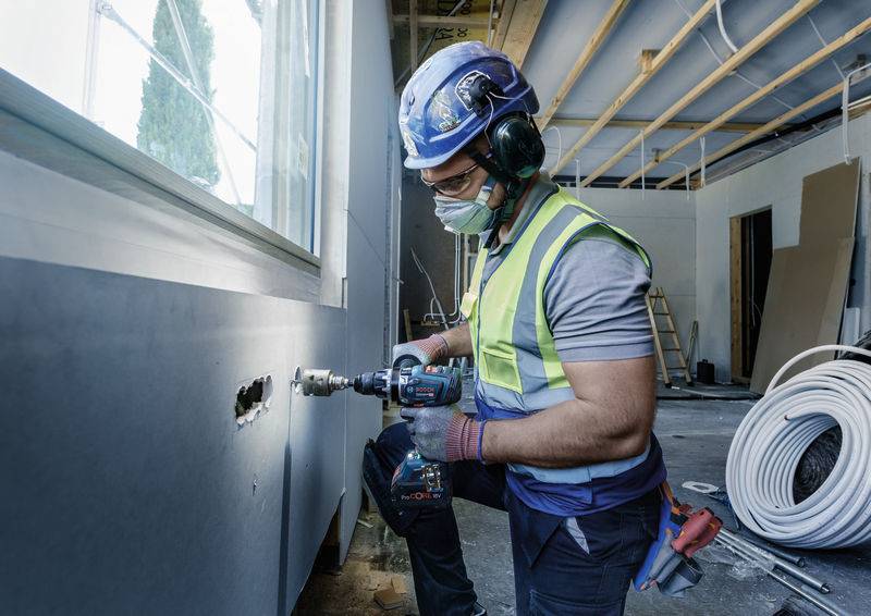 Dělník v ochranném oděvu a helmě vrtá díru do zdi v nedokončené místnosti; probíhá stavba a renovace.