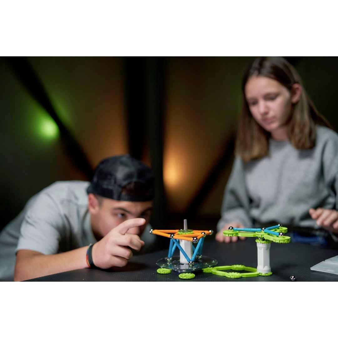 Dvě osoby sedí u stolu a staví model s barevnými díly, soustředěně a s nadšením při práci.