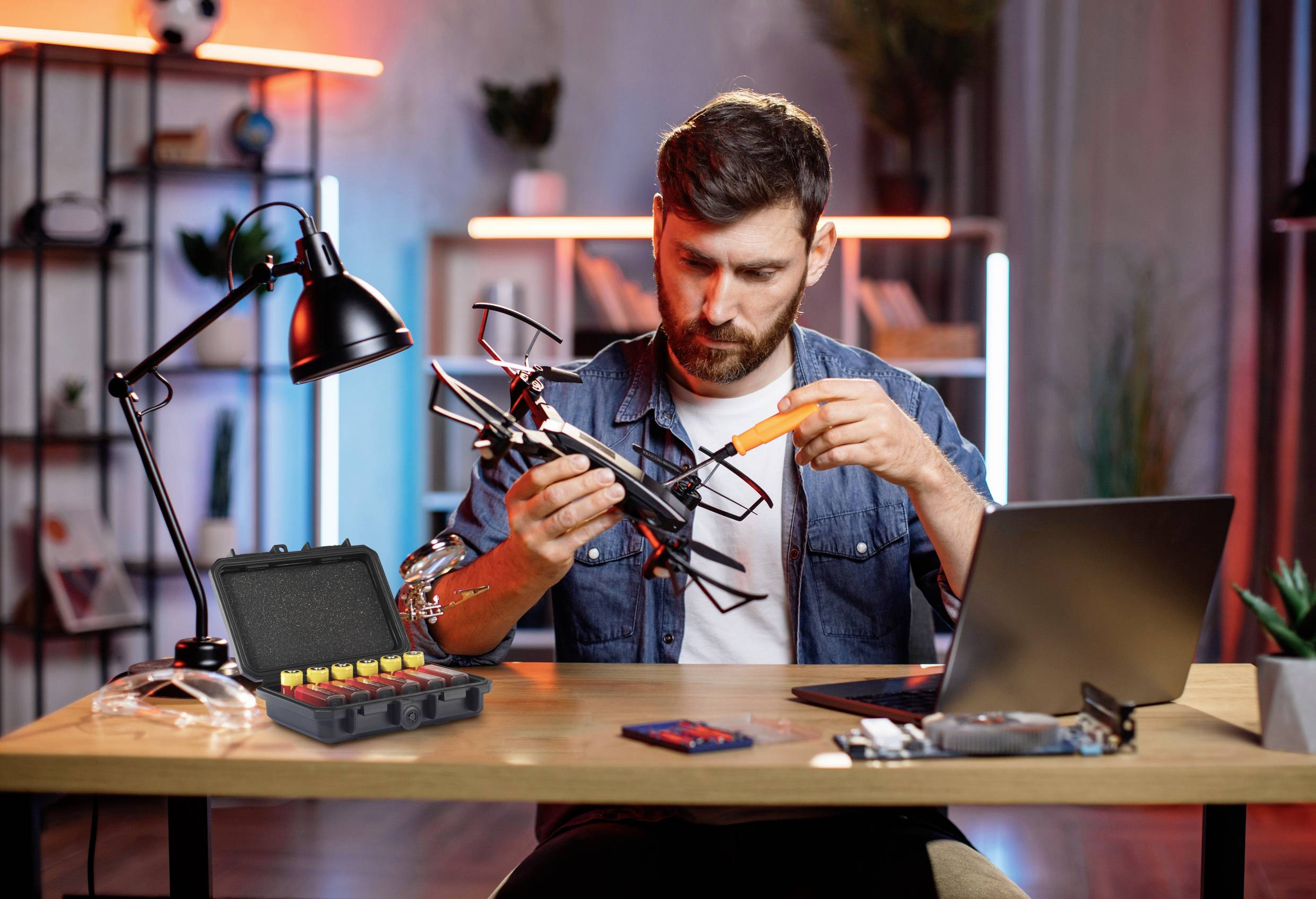 Muž sedí u stolu a opravuje dron. Vedle něj leží laptop a otevřený kufřík s nástroji a bateriemi.