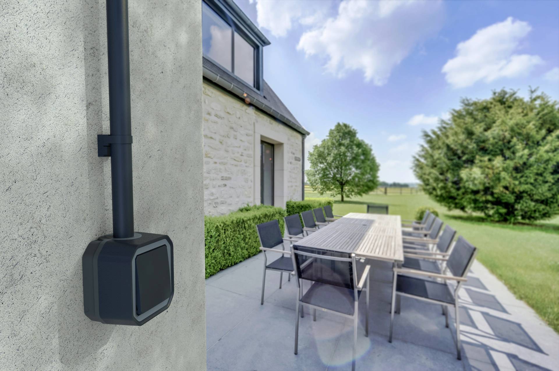 Slunečná zahrada s dlouhým stolem a židlemi na terase, obklopená udržovaným trávníkem a stromy; moderní dům v pozadí.