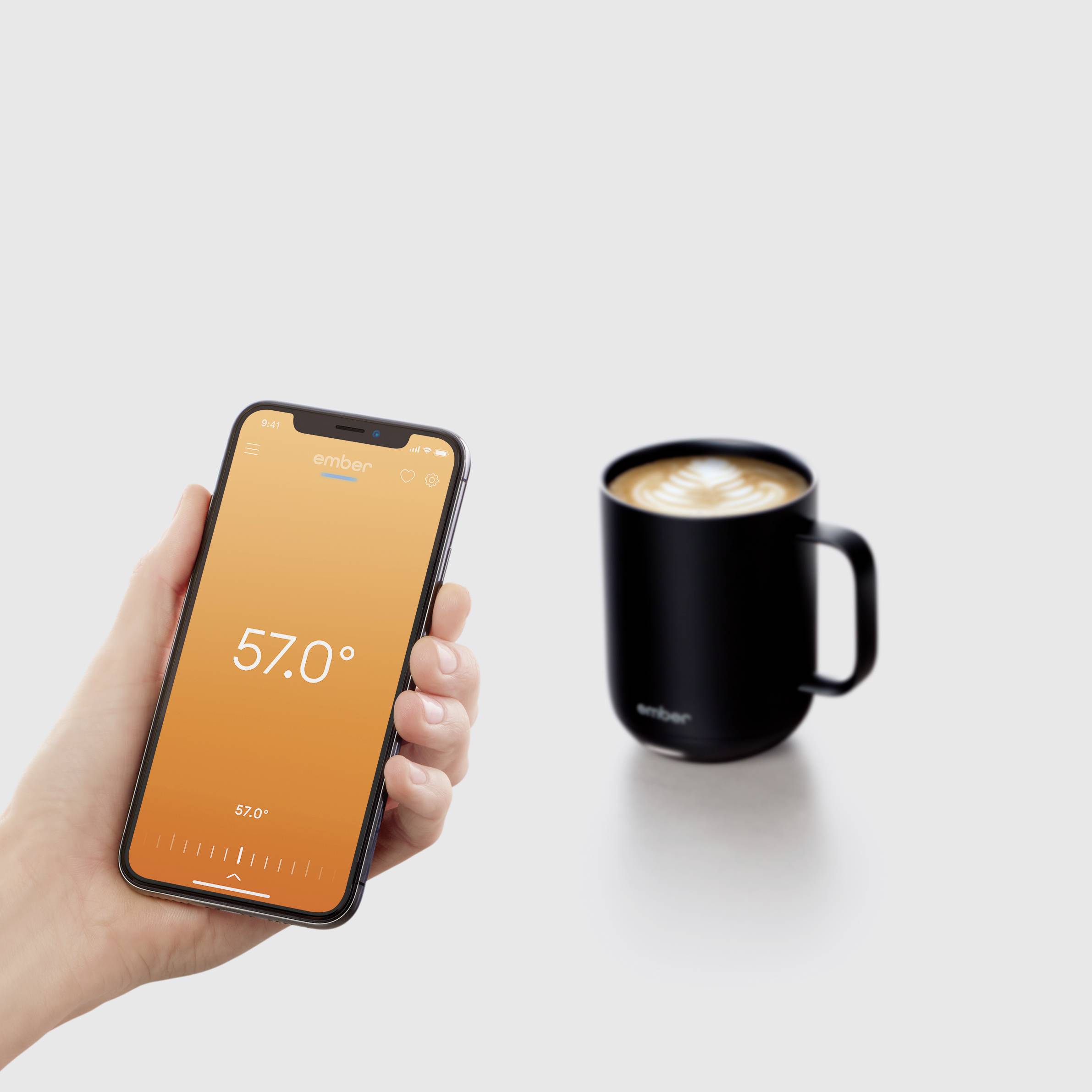 Ruka drží smartphone s aplikací zobrazující 57,0 stupňů. Vedle stojí černý šálek s kávou a latte art.