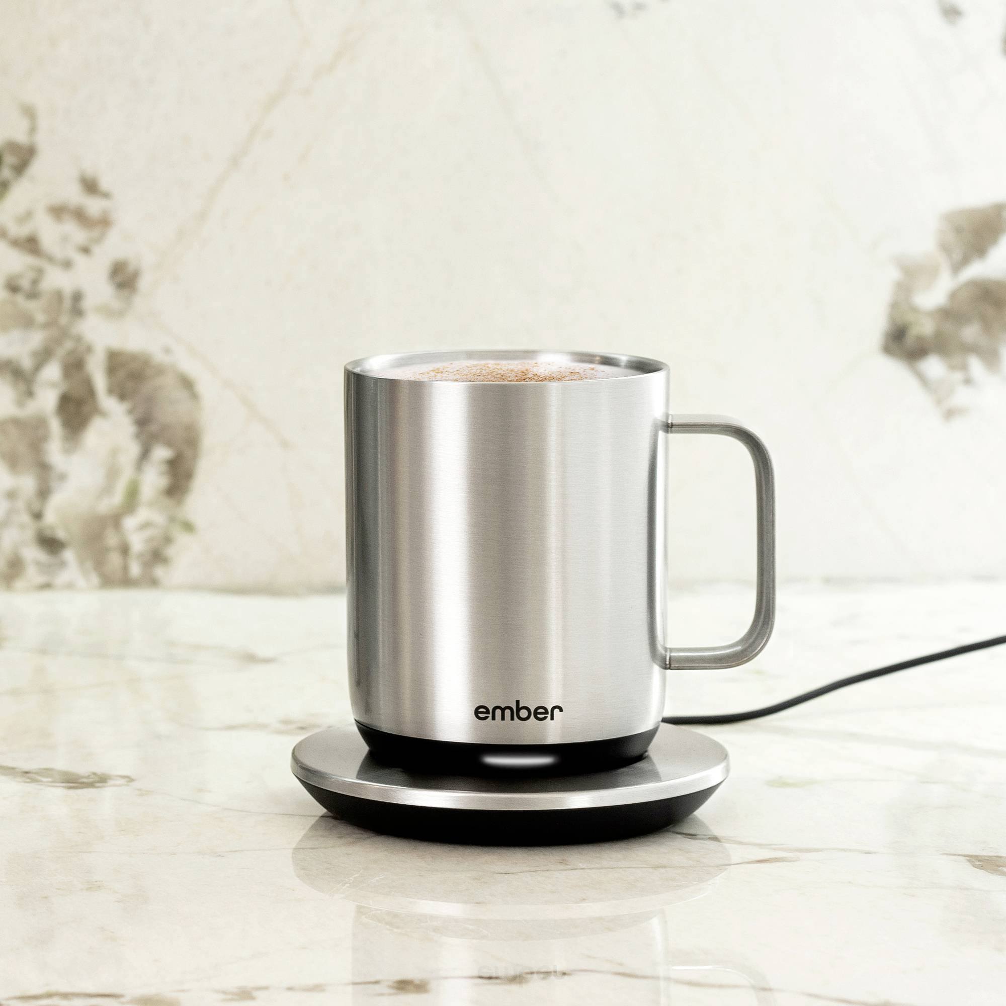 Stříbrný hrnek s potiskem 'ember' stojí na nabíjecí stanici, naplněný horkým nápojem. Nachází se na mramorové ploše.