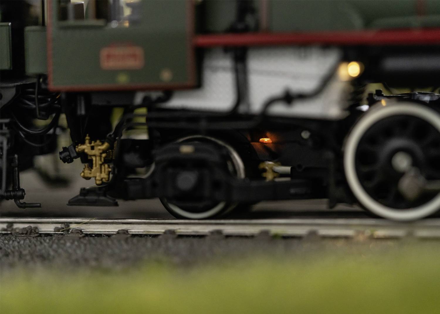 Detailní záběr modelové železnice na kolejích. V popředí jsou viditelná kola a mechanické součásti. Pozadí je rozostřené.