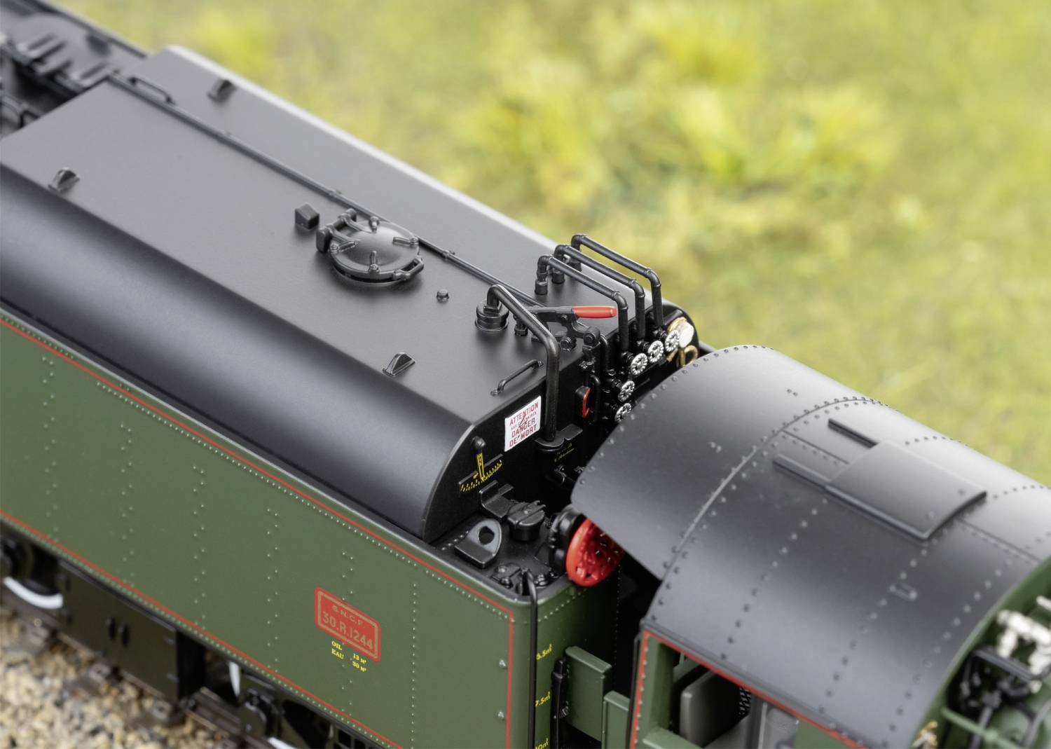 Detailní záběr modelové lokomotivy v zelené barvě s černými a červenými akcenty, umístěné na kolejích s rozostřeným travnatým pozadím.