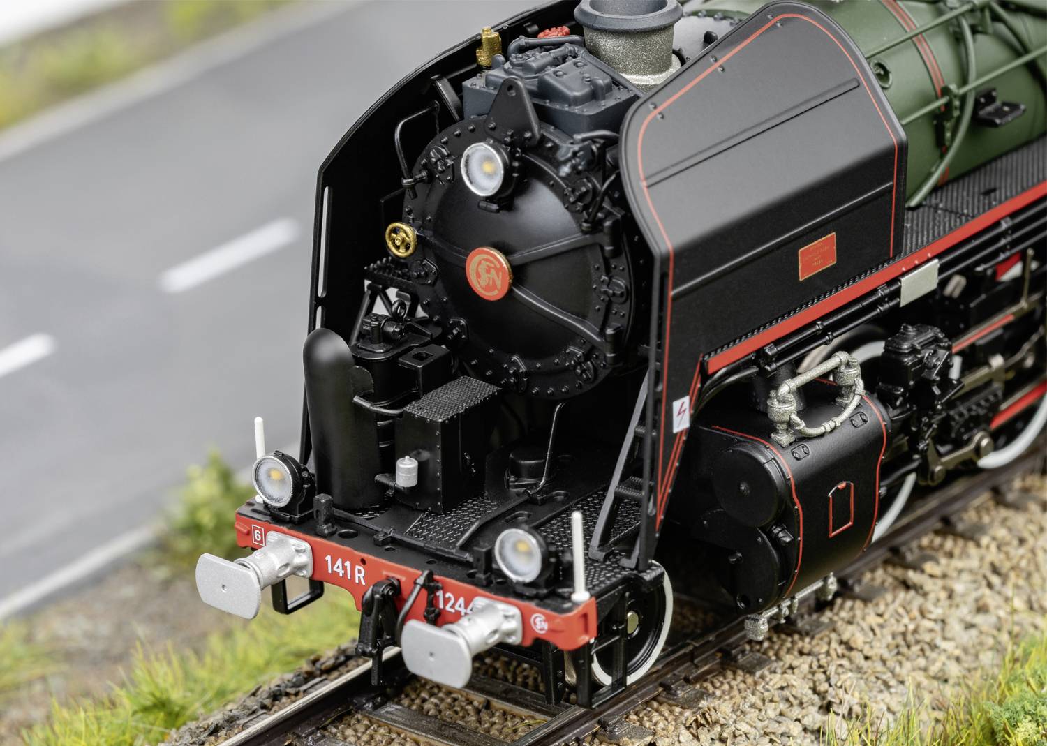 Detailní model parní lokomotivy na kolejích vedle silnice, který ukazuje čelní pohled s detaily kotle.