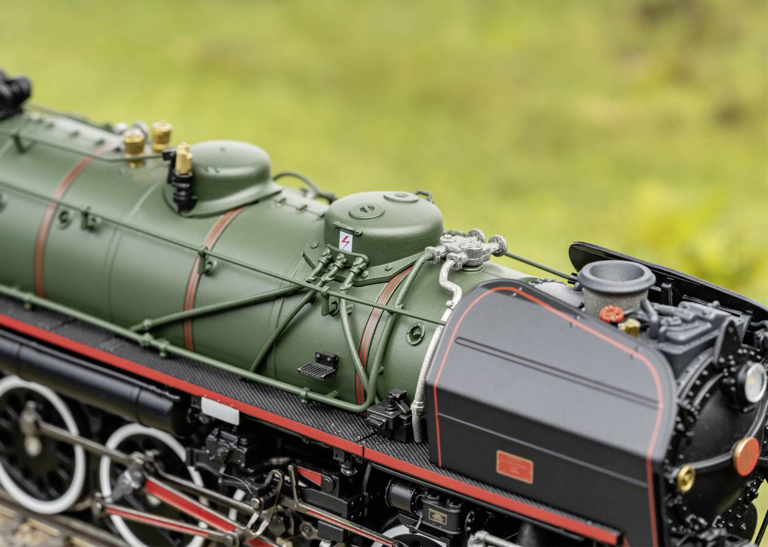 Model historické parní lokomotivy se zeleným kotlem, černou kabinou strojvedoucího, červenými detaily a stříbrnými ventily v popředí.