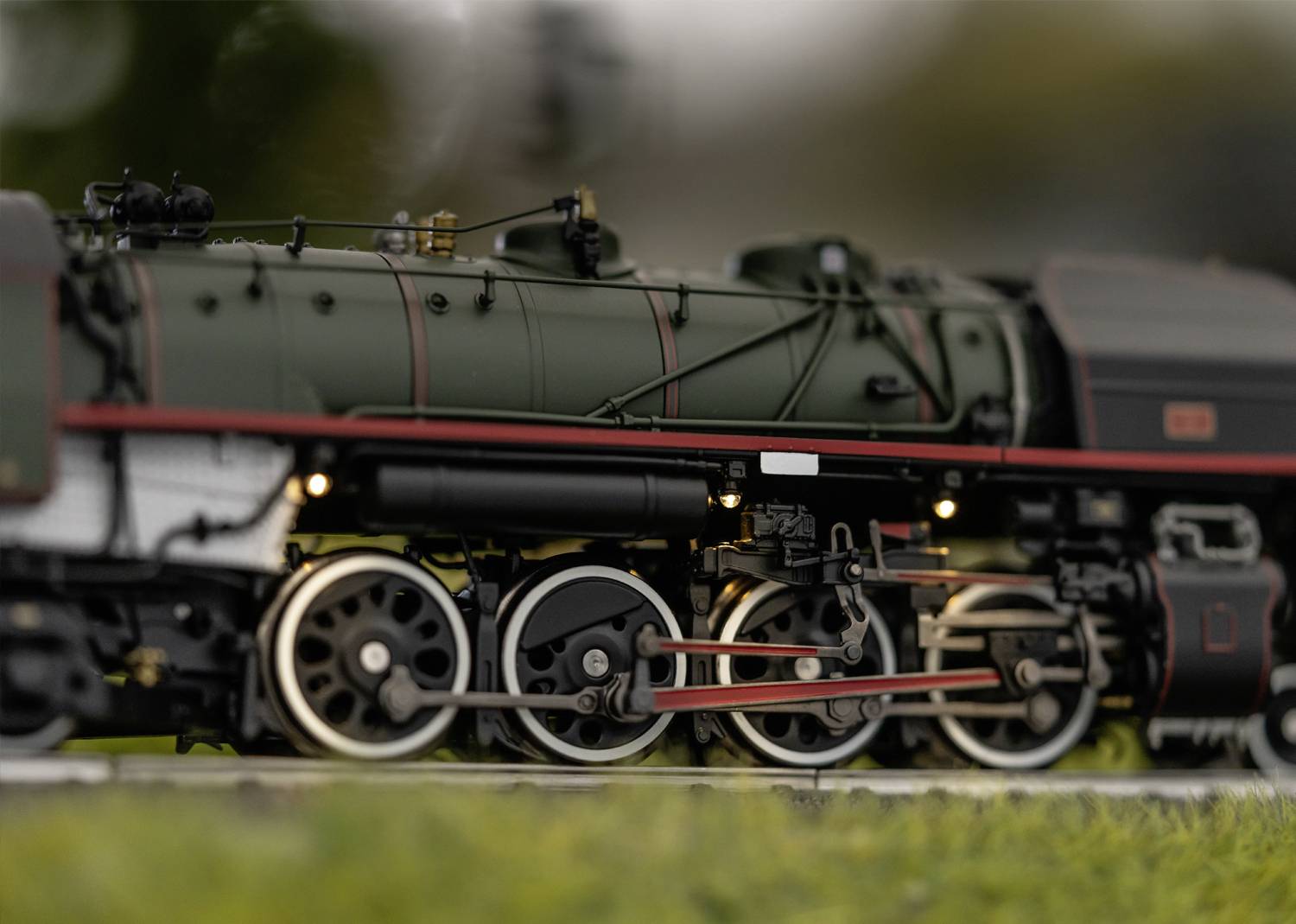 Detailní modelová lokomotiva na kolejích, obklopená rozostřenou zelení v popředí i pozadí. Vášeň pro techniku.