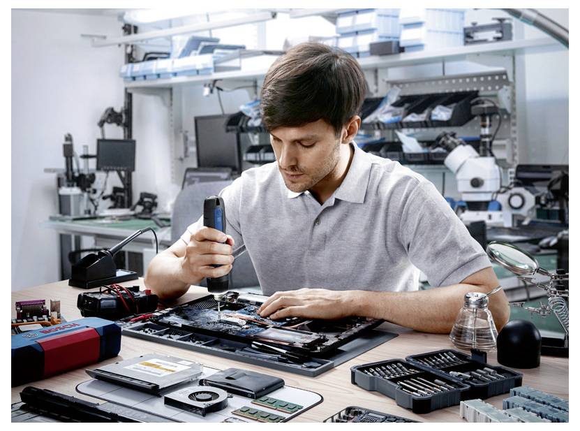 Muž opravuje elektronické zařízení na stole, obklopen nástroji a náhradními díly před laboratorním pozadím.