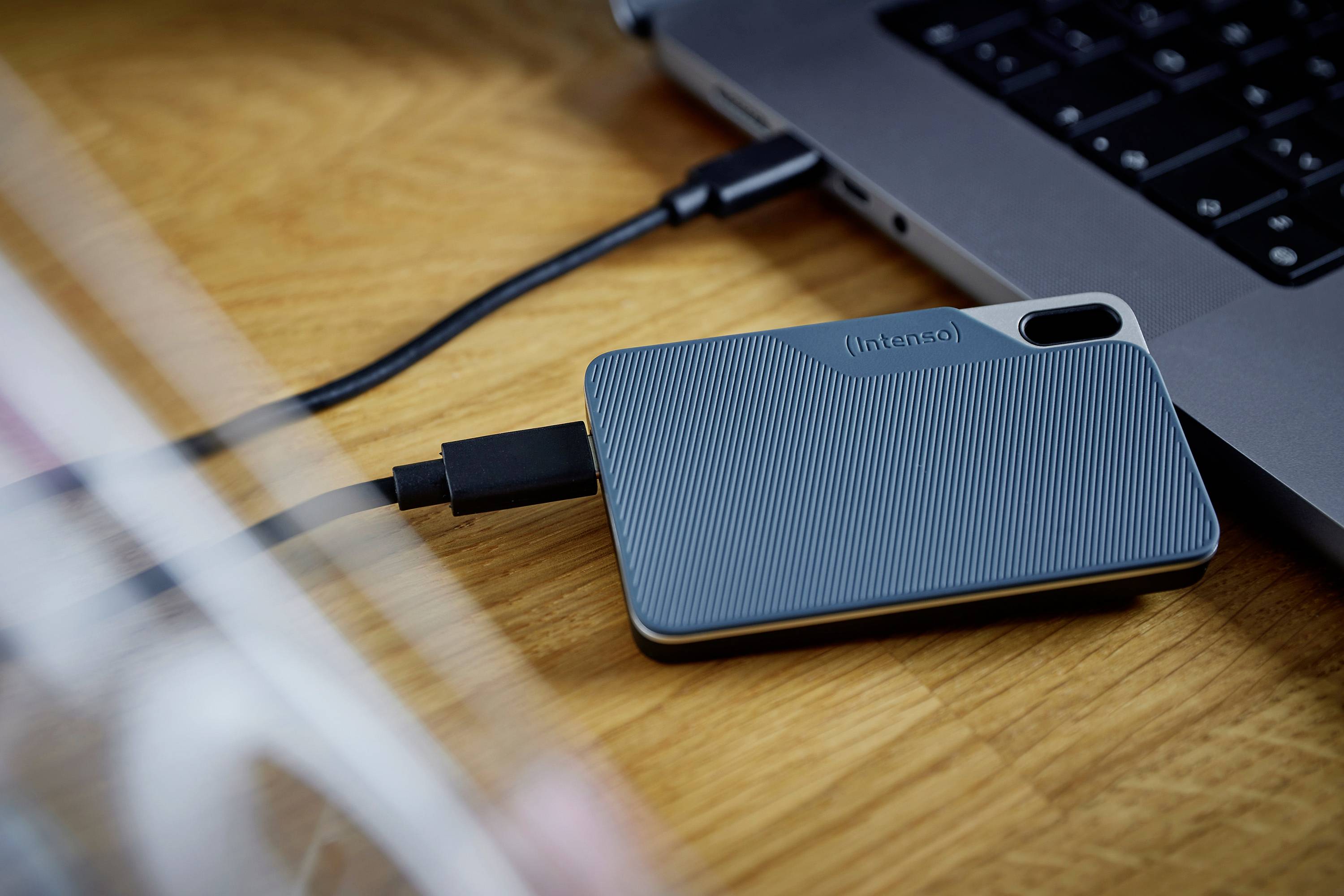 Modrý externí pevný disk připojený k laptopu na dřevěném stole. Pevný disk slouží k ukládání dat.