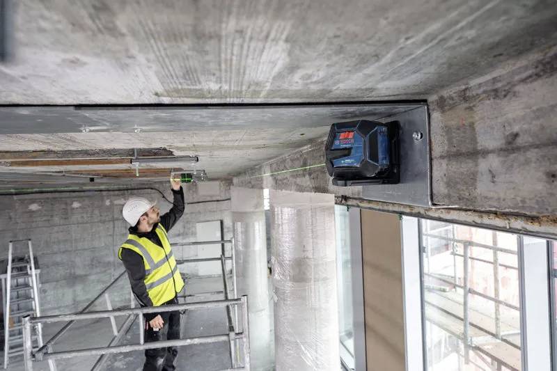 Stavební dělník v helmě a reflexní vestě měří vzdálenost na betonové stropní desce pomocí přesného měřicího nástroje na staveništi.