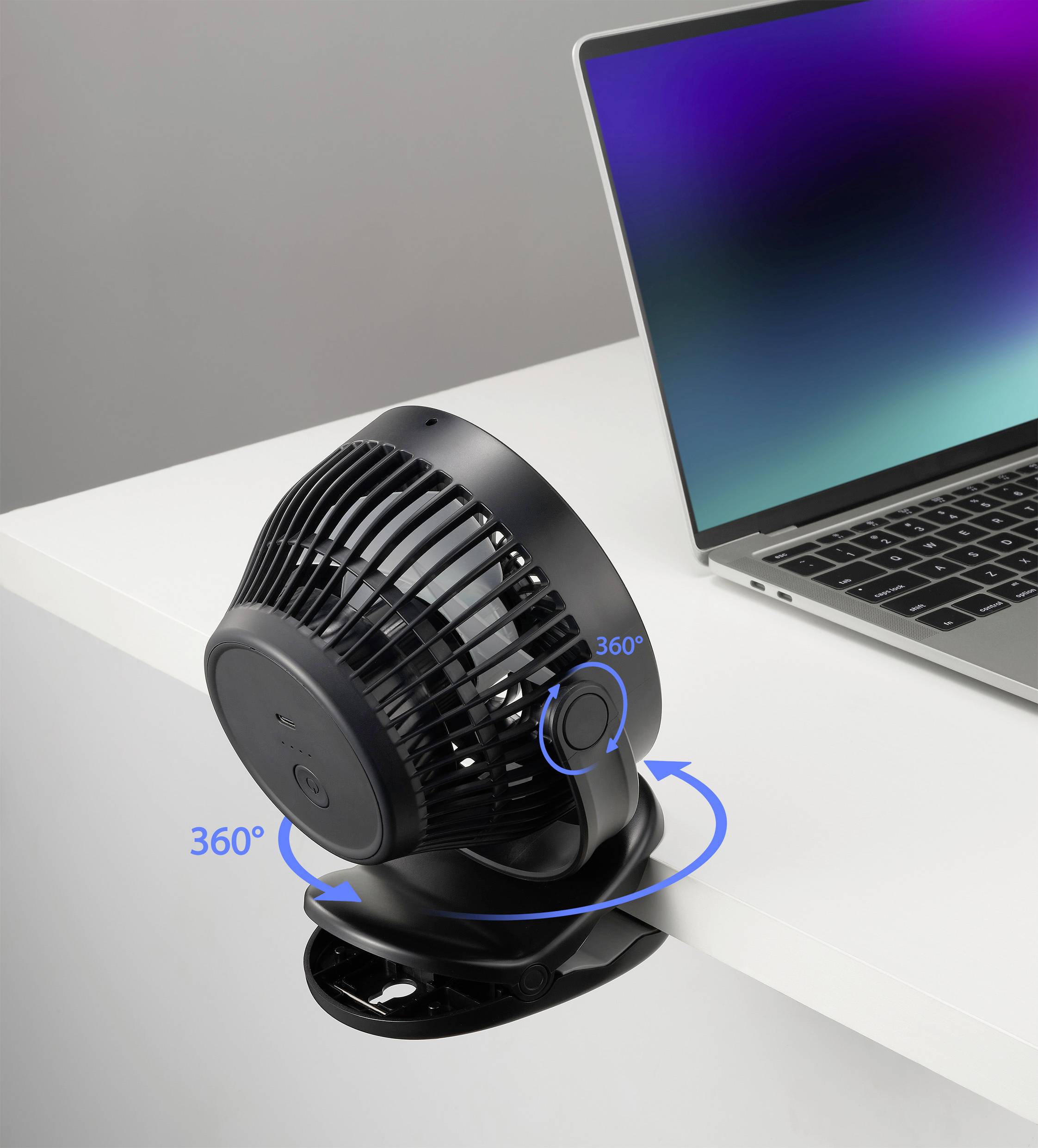 Černý, kulatý ventilátor je připevněn na okraji stolu a může se otáčet o 360 stupňů. Vedle něj stojí laptop.