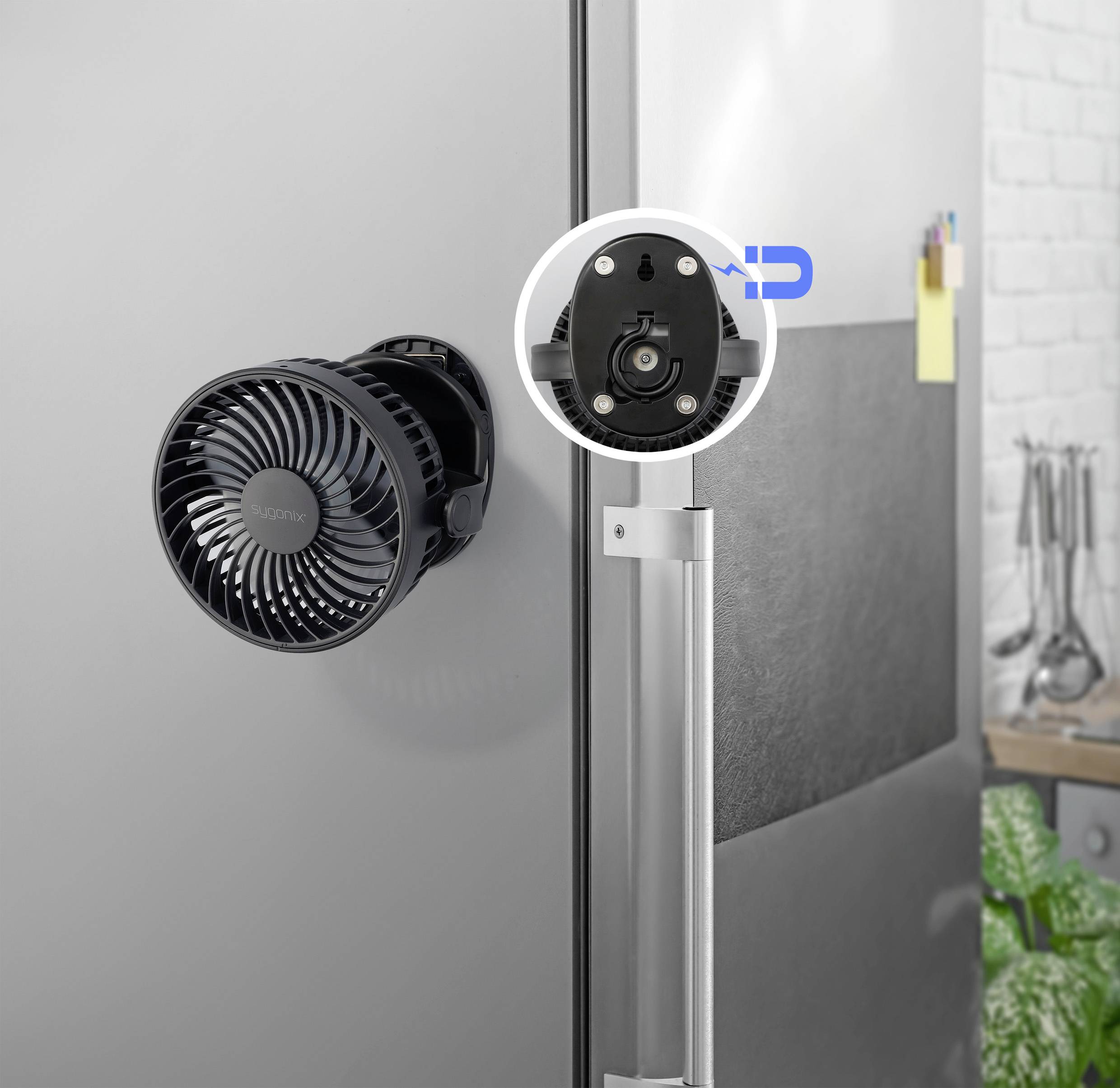 Černý ventilátor je připevněn pomocí magnetu na dveře lednice. V pozadí kuchyně jsou vidět různé kuchyňské pomůcky.