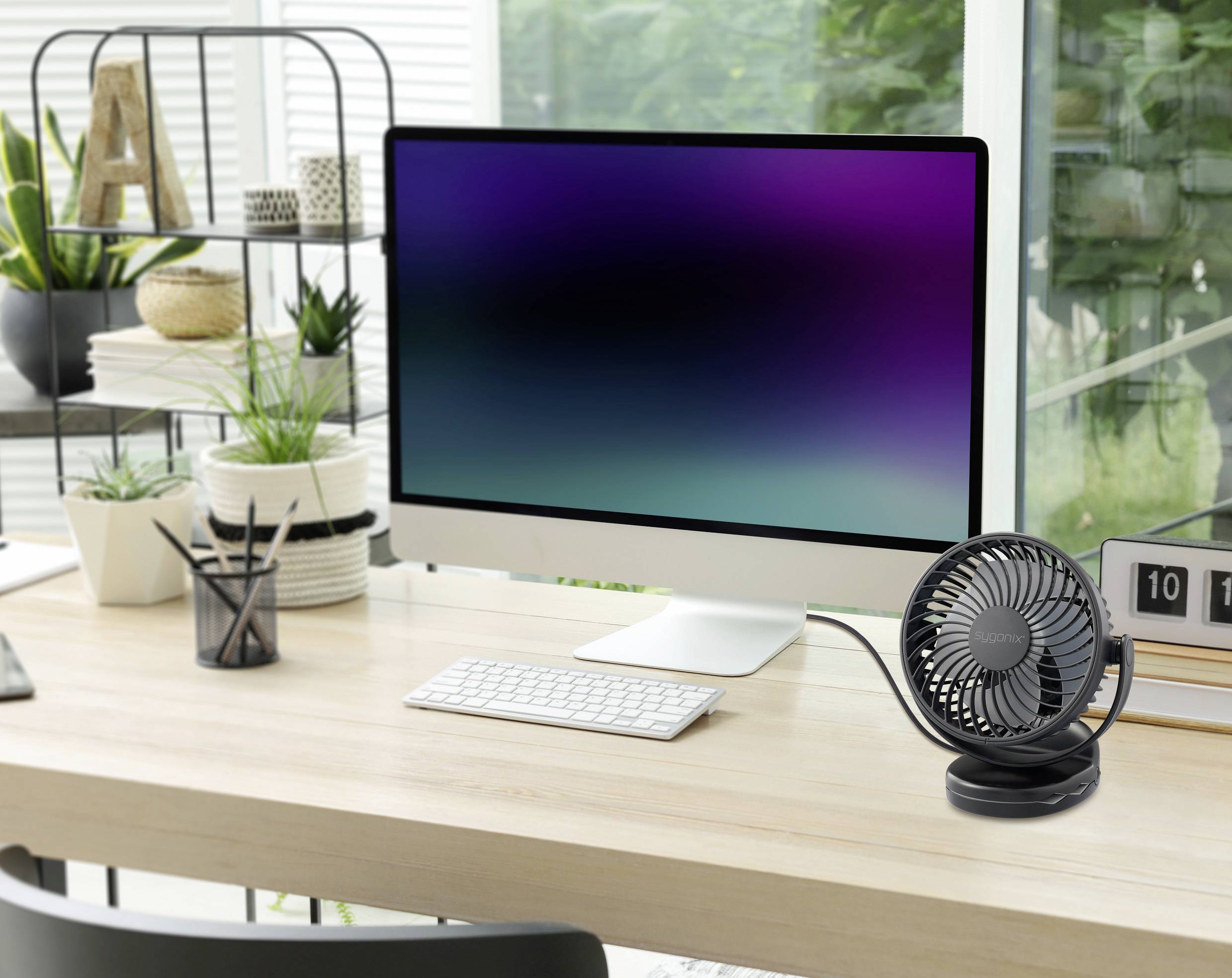 Moderní psací stůl s velkým počítačovým monitorem, bezdrátovou klávesnicí a myší. Vpravo stojí malý ventilátor, obklopený rostlinami.