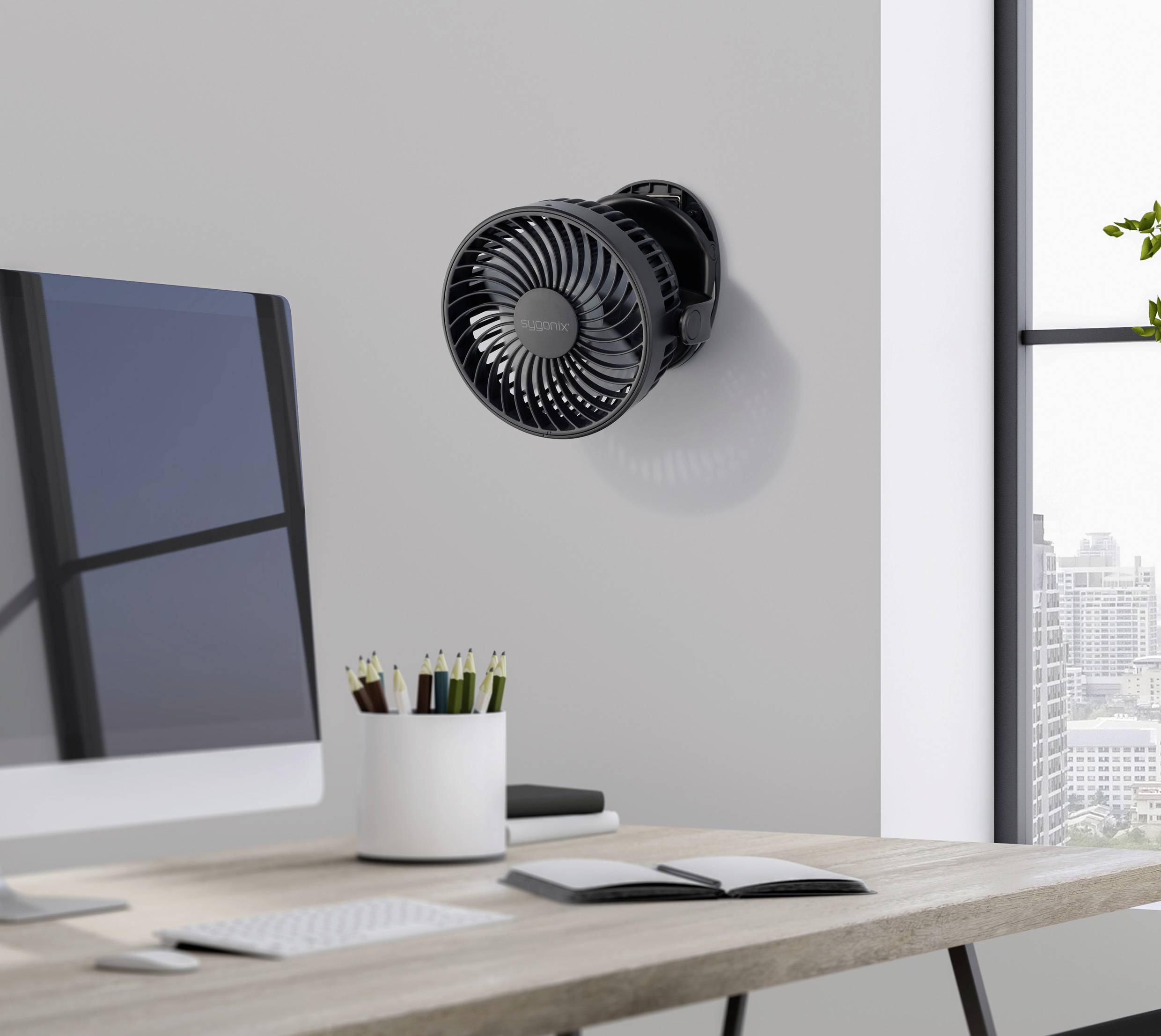 Černý ventilátor je namontován na stěně moderní kanceláře. Místnost obsahuje psací stůl s kancelářskými potřebami a okno s výhledem na město.