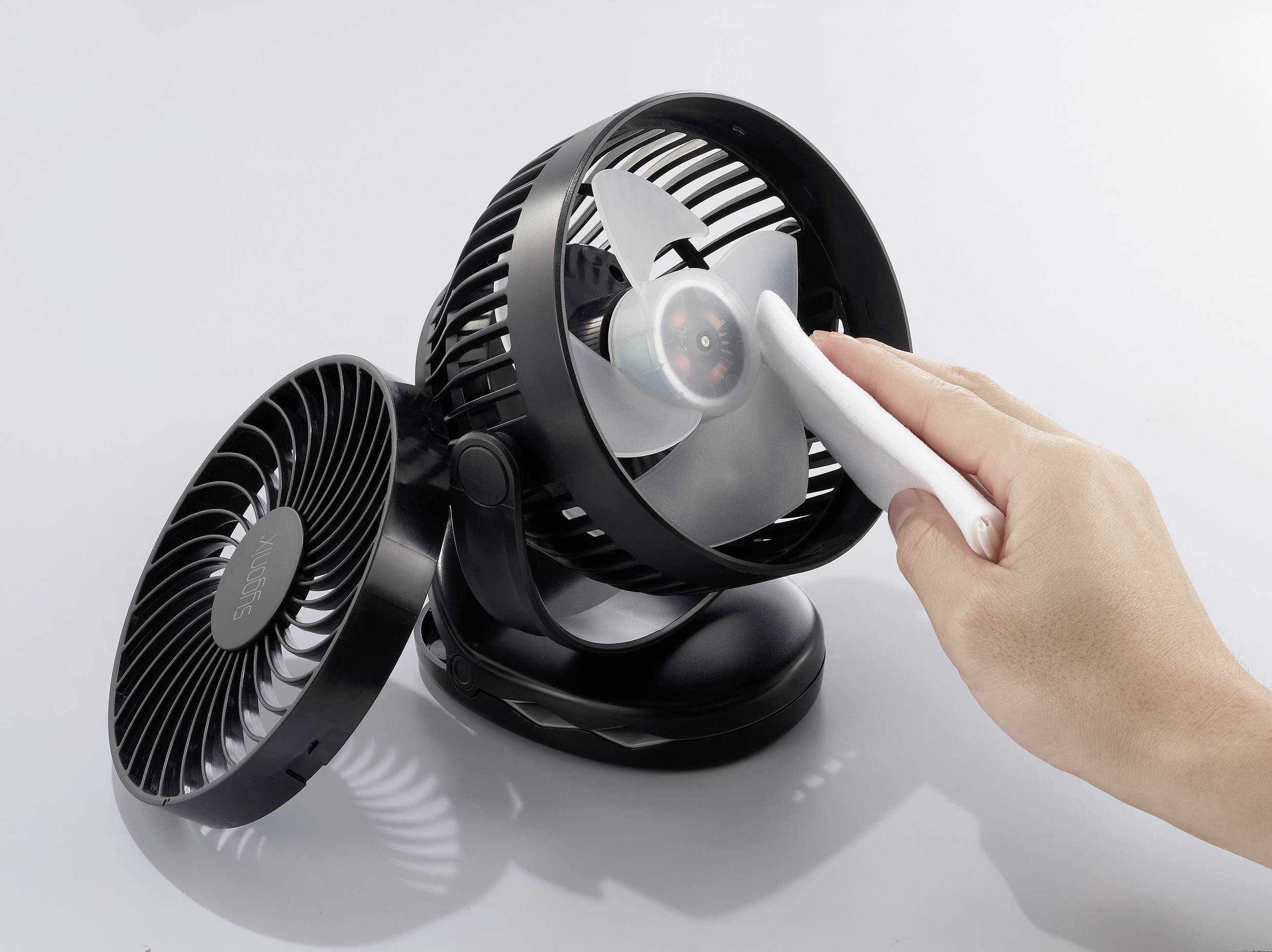 Ruka čistí lopatky malého černošedého ventilátoru bílým kartáčem. V pozadí je druhý ventilátor.