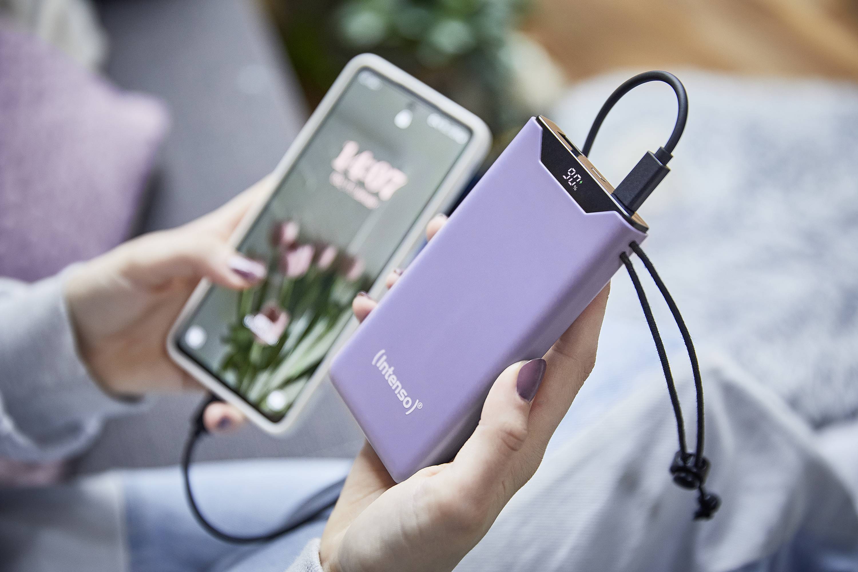 Osoba drží smartphone s květinovým pozadím. Vedle je fialová powerbanka, která nabíjí telefon.