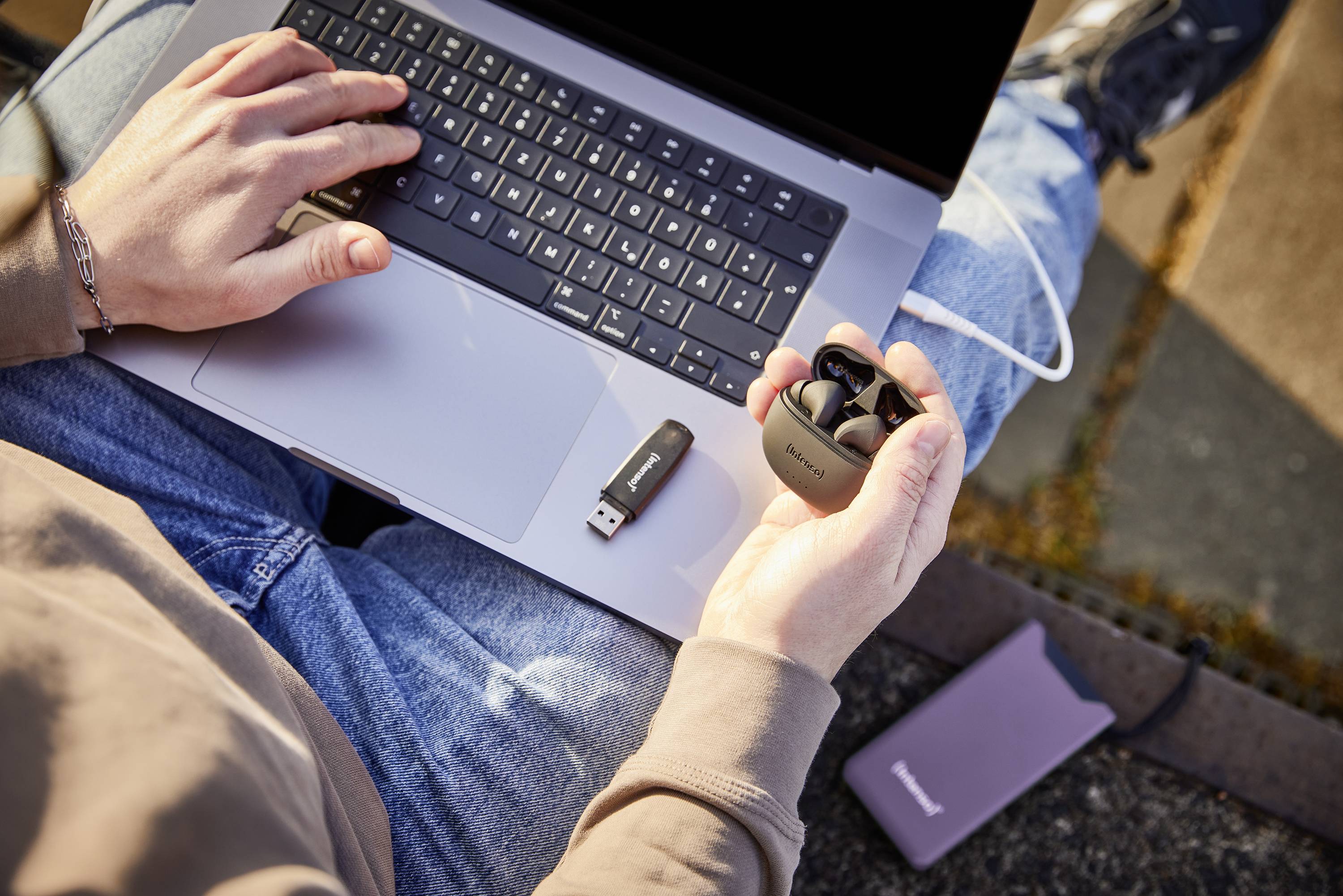 Osoba sedí venku s laptopem na klíně, drží bezdrátová sluchátka v ruce a vedle má položený USB flash disk.
