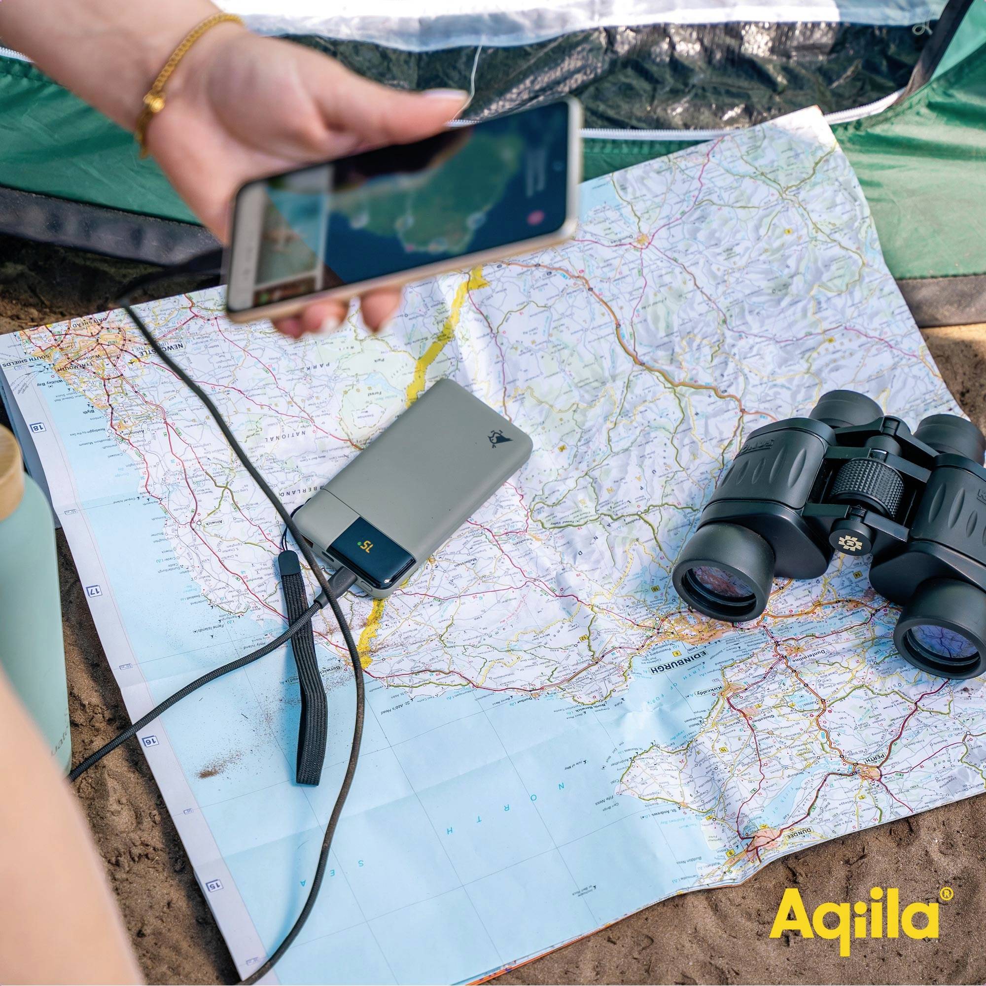 Osoba se dívá na smartphone nad mapou ležící na zemi vedle páru dalekohledů, s viditelnou nálepkou 'Aqiila'.