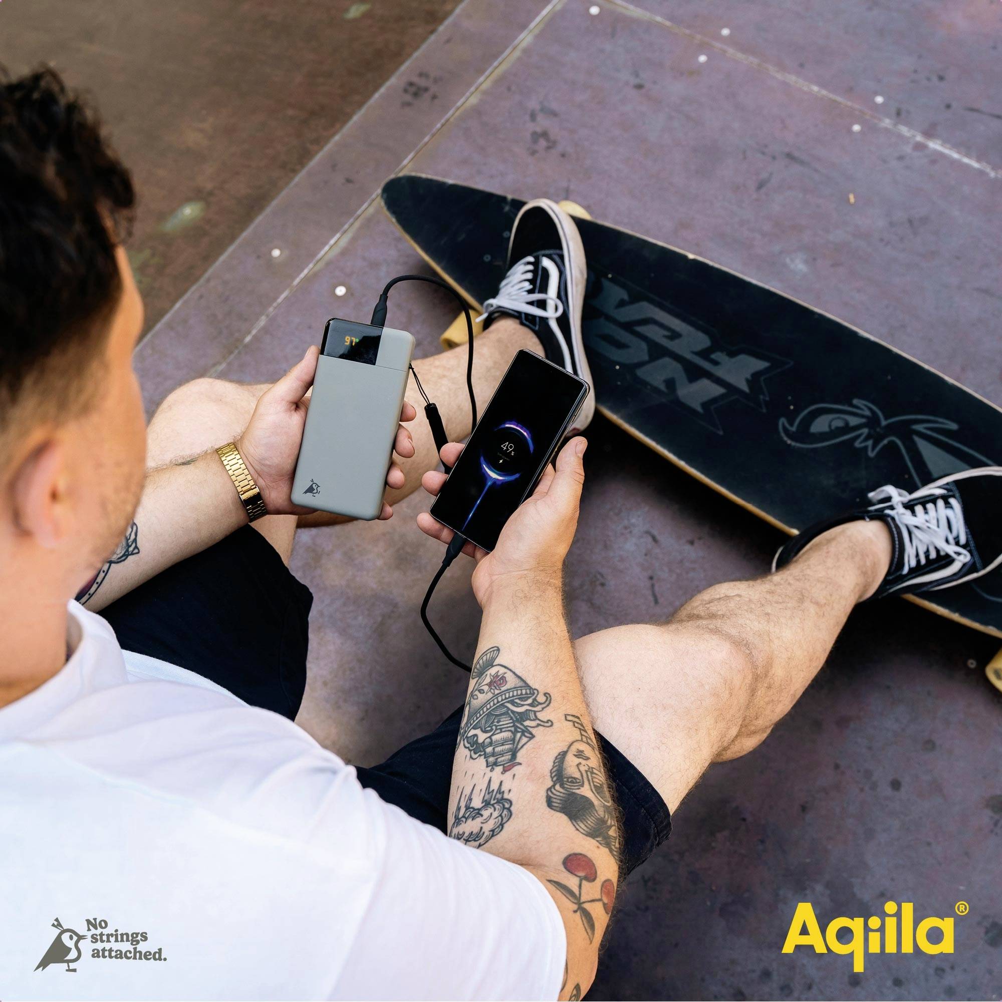Muž sedí na skateboardu a drží v ruce smartphone připojený k přenosnému powerbance.