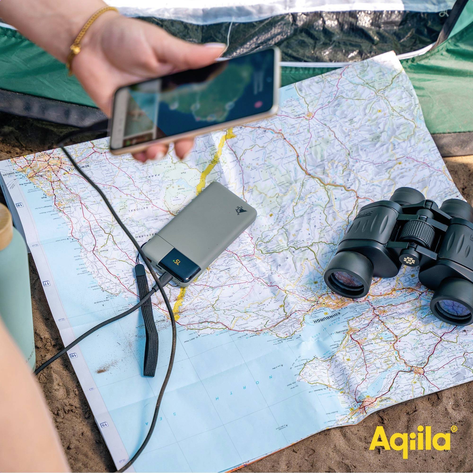 Na zemi leží mapa a na ní smartphone, dalekohled a nabíječka. Jedna osoba drží další smartphone.