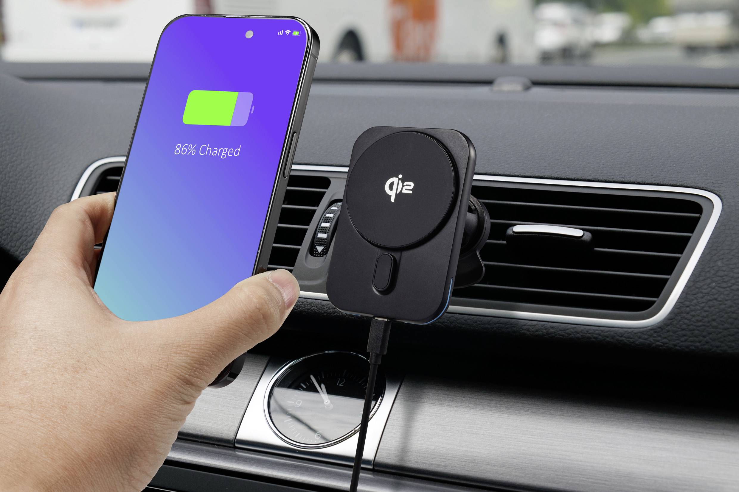 Jedna ruka drží smartphone s obrazovkou ukazující '86% nabito' před bezdrátovou Qi nabíječkou připevněnou k ventilační mřížce v autě.