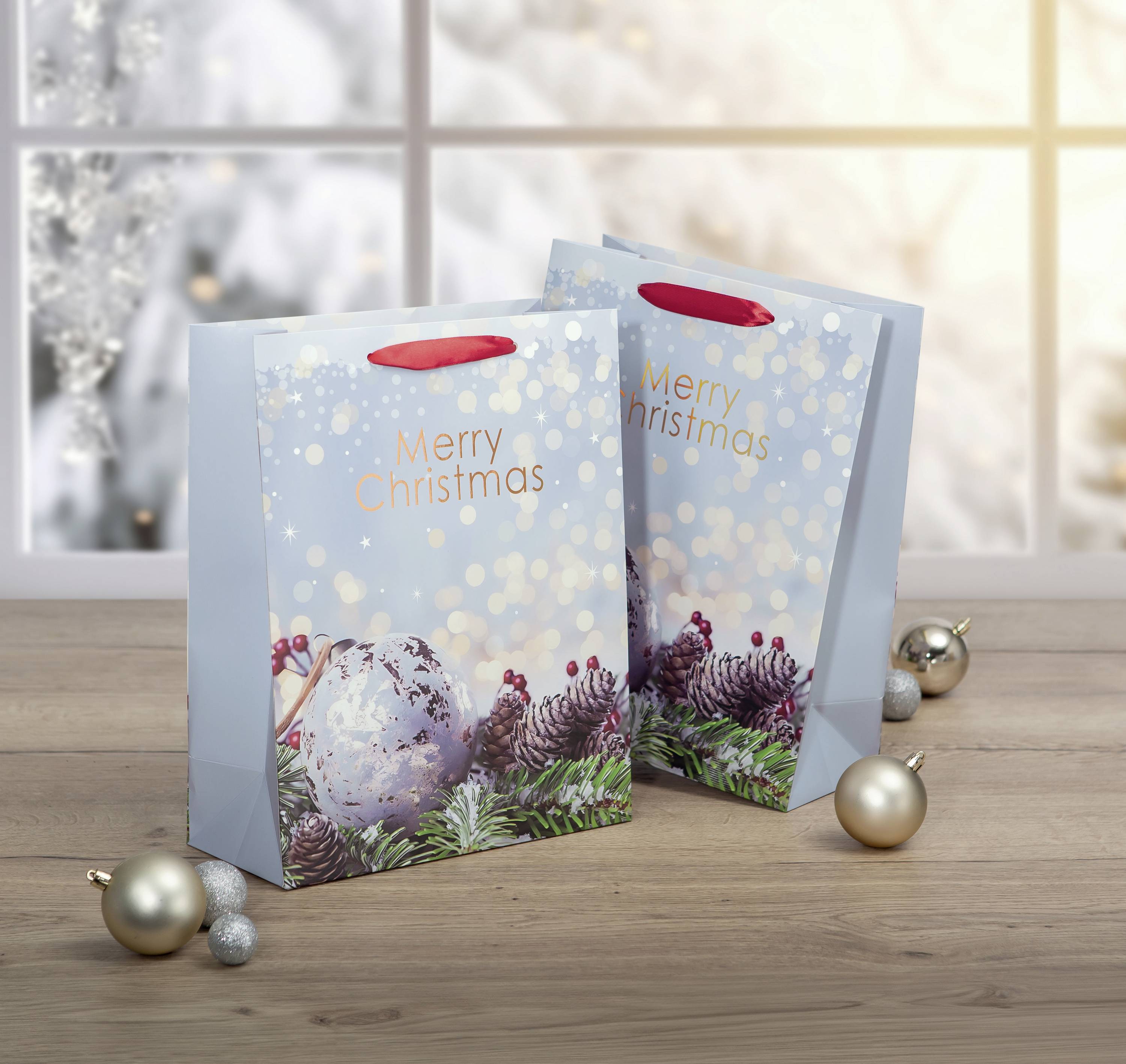 Dvě vánočně ozdobené dárkové tašky s nápisem 'Merry Christmas' stojí na dřevěném stole, obklopené stříbrnými koulemi.
