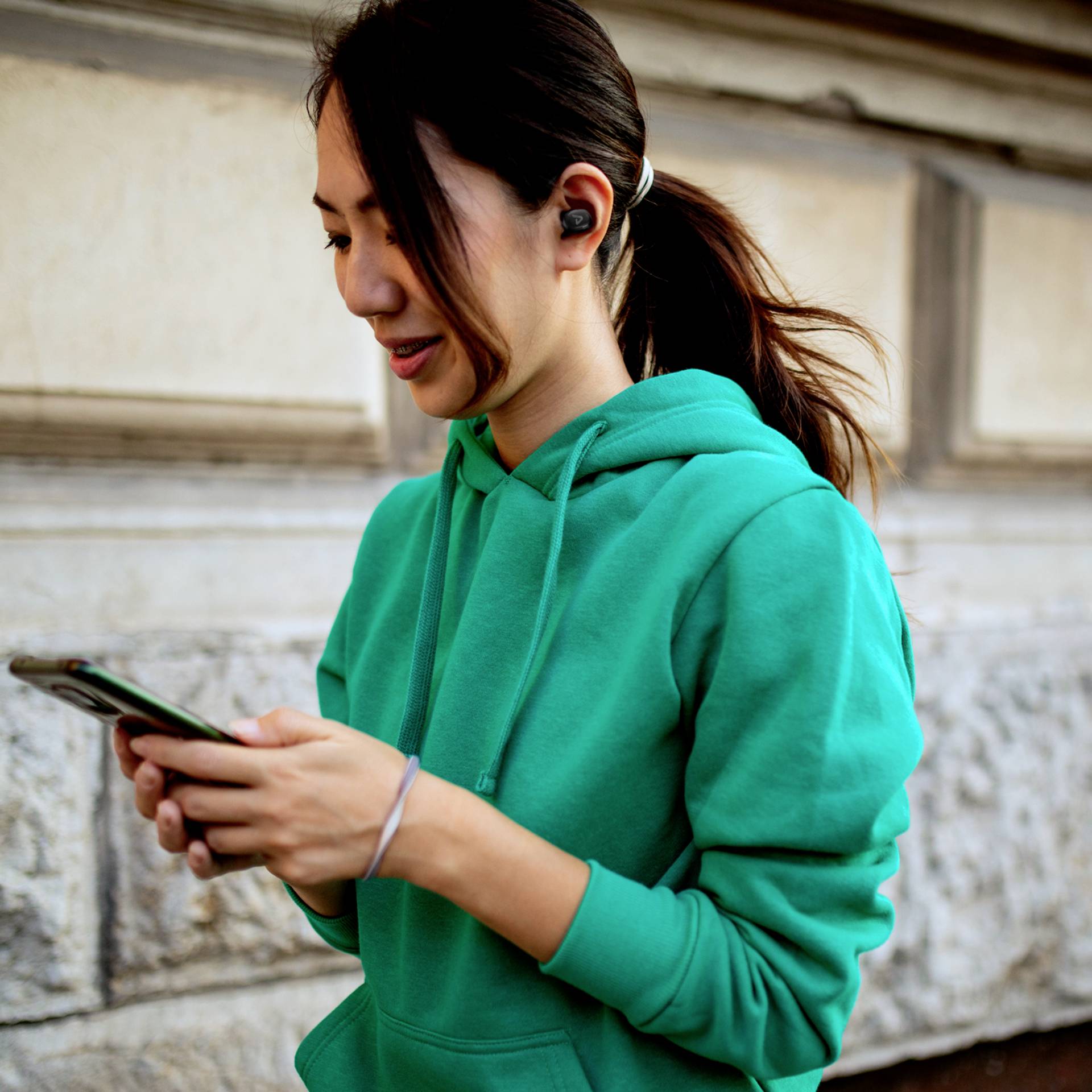 Usměvavá osoba v zeleném mikině stojí venku a dívá se na svůj smartphone, přičemž má nasazená bezdrátová sluchátka.