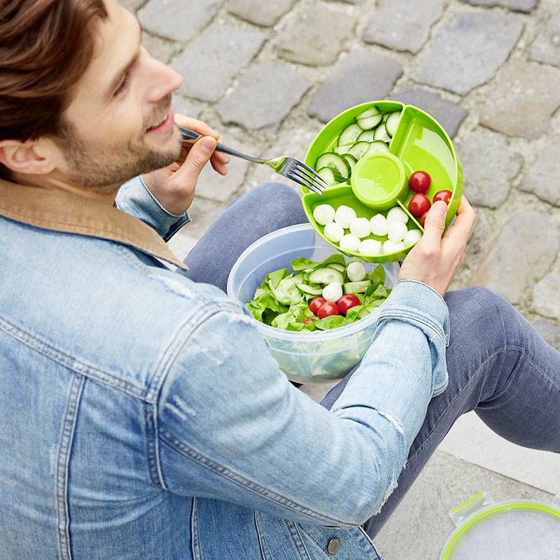 Muž sedí venku na kamenné zdi a jí salát z kulatého plastového boxu s přihrádkami pro různé ingredience.