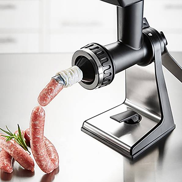 Kuchyňský robot tvaruje syrové mleté maso do párků. Čerstvé rozmarýnové větvičky leží vedle na kovovém stole.