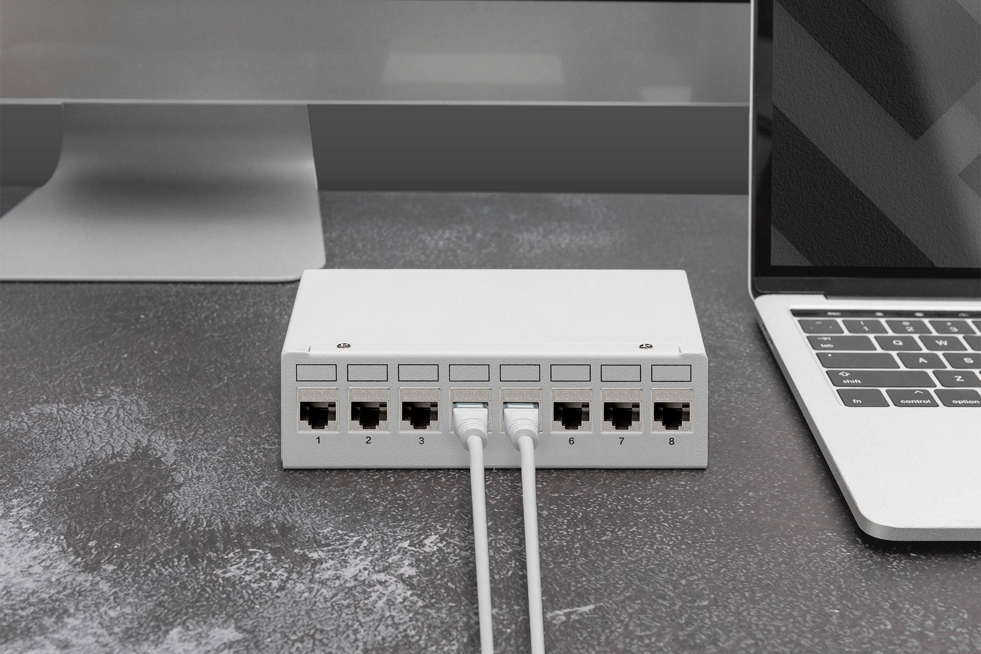 Bílý síťový switch s osmi ethernetovými porty stojí na stole, propojený dvěma šedými kabely. Vedle něj se nachází monitor a laptop.