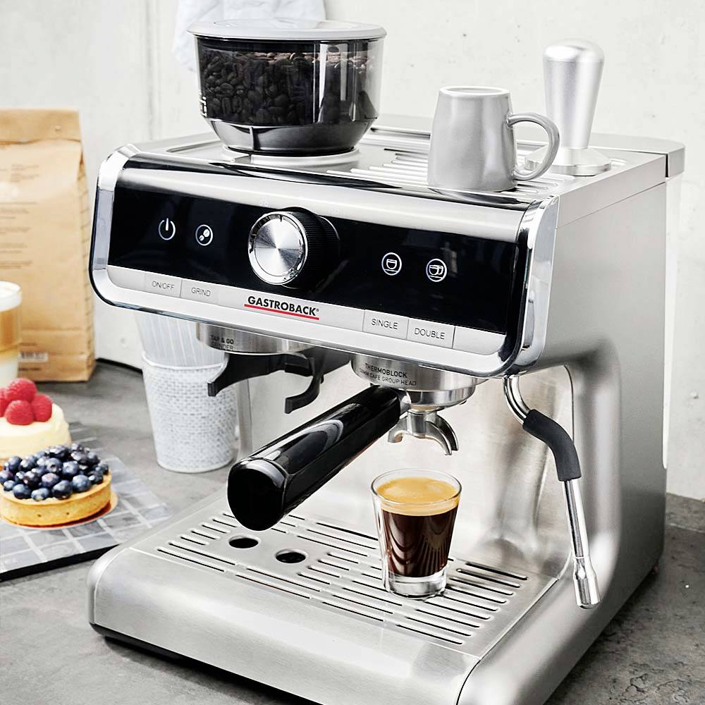 Stříbrný espresso přístroj právě připravuje espresso. Nádoba na kávová zrna je umístěna na stroji. V pozadí je dort.
