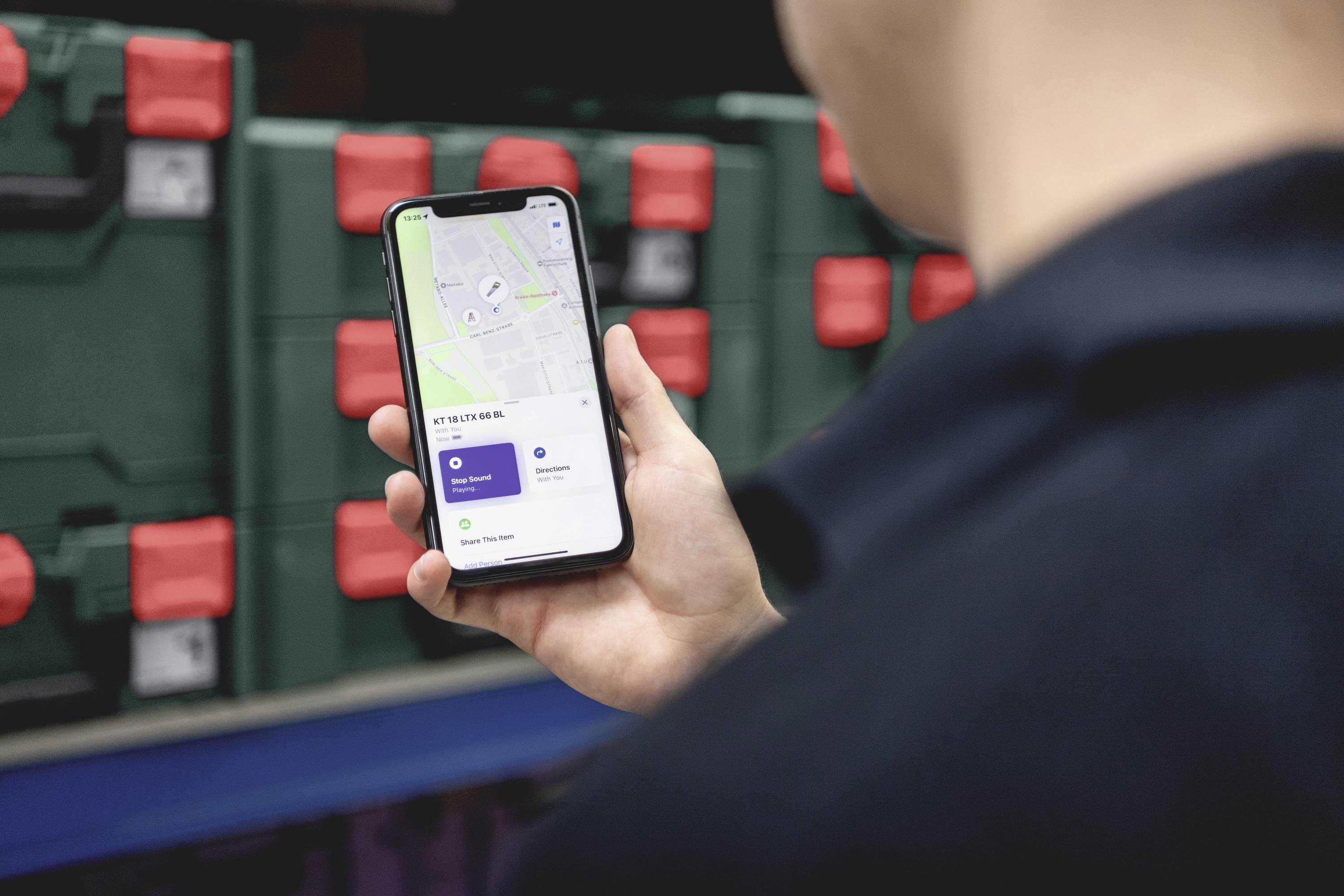 Osoba drží smartphone s otevřenou navigační aplikací. V pozadí jsou vidět zelené bedny s červenými uzávěry.