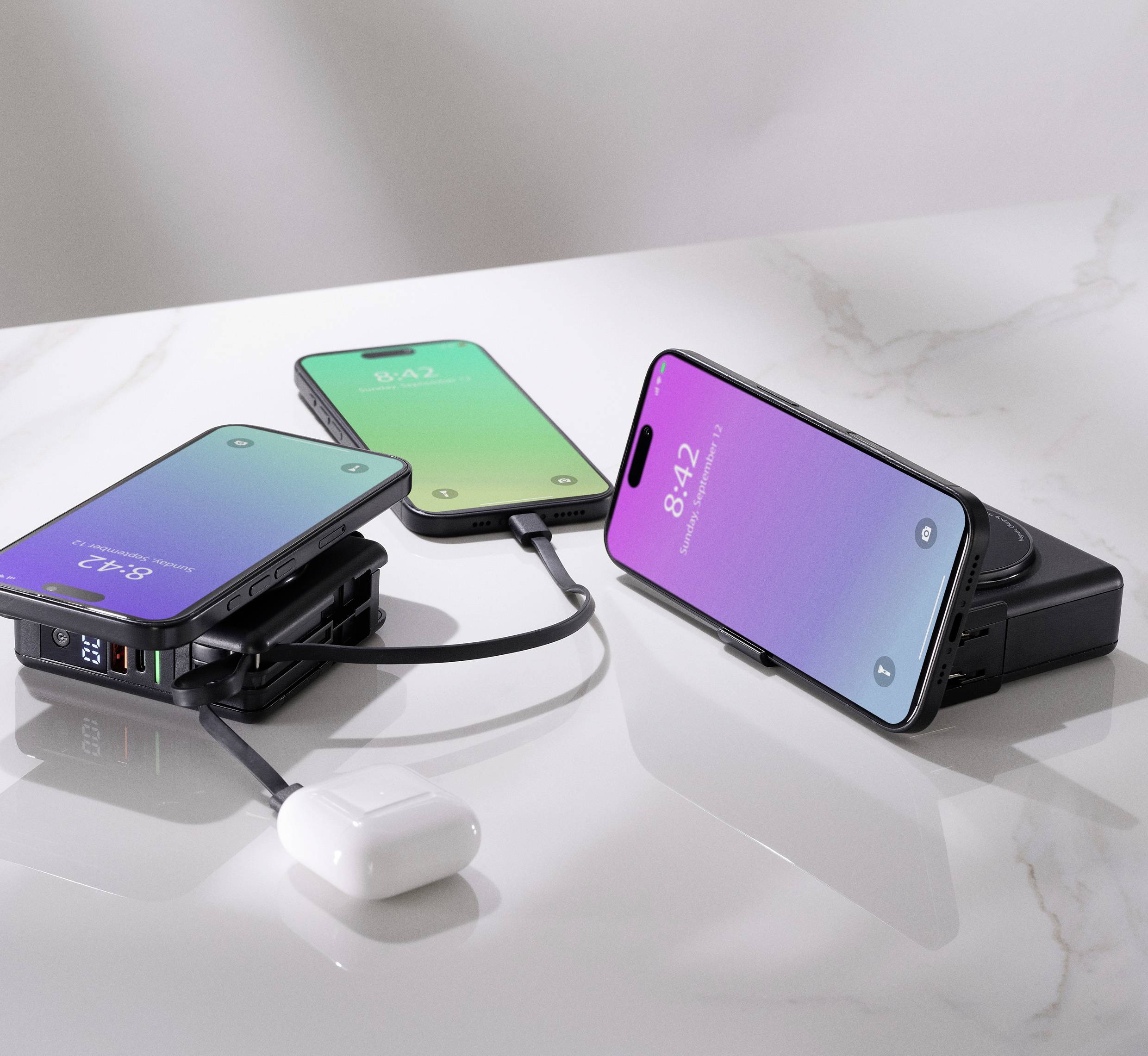 Tři smartphony a bezdrátová sluchátka na stole, dva telefony se nabíjejí pomocí kabelů a třetí je umístěn na bezdrátové nabíječce.