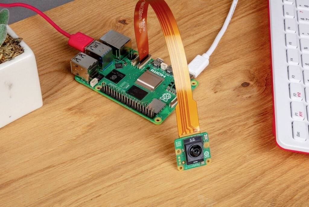 Deska Raspberry Pi s připojeným kamerovým modulem ležící na dřevěném stole ukazuje kompaktní počítačové zařízení.