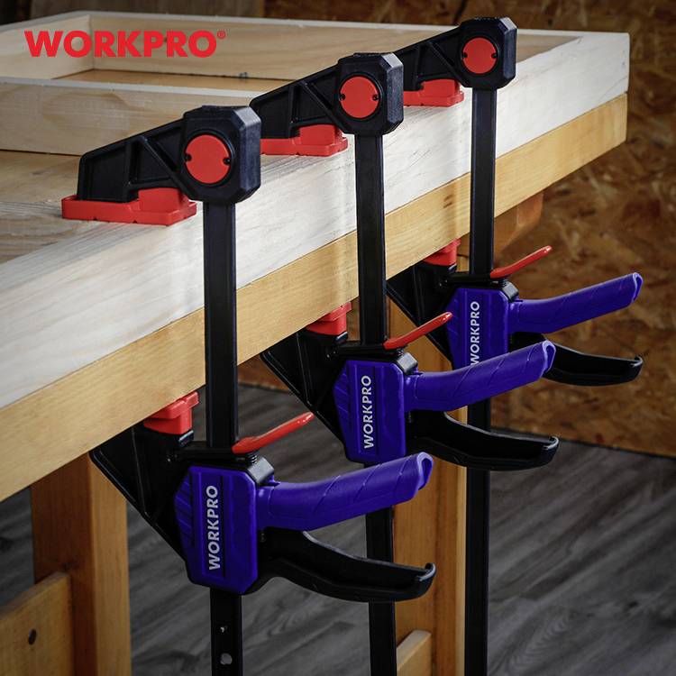 Tři černé a červené svěrky drží dřevěnou desku na stole. Na svěrkách je viditelný nápis "WORKPRO".
