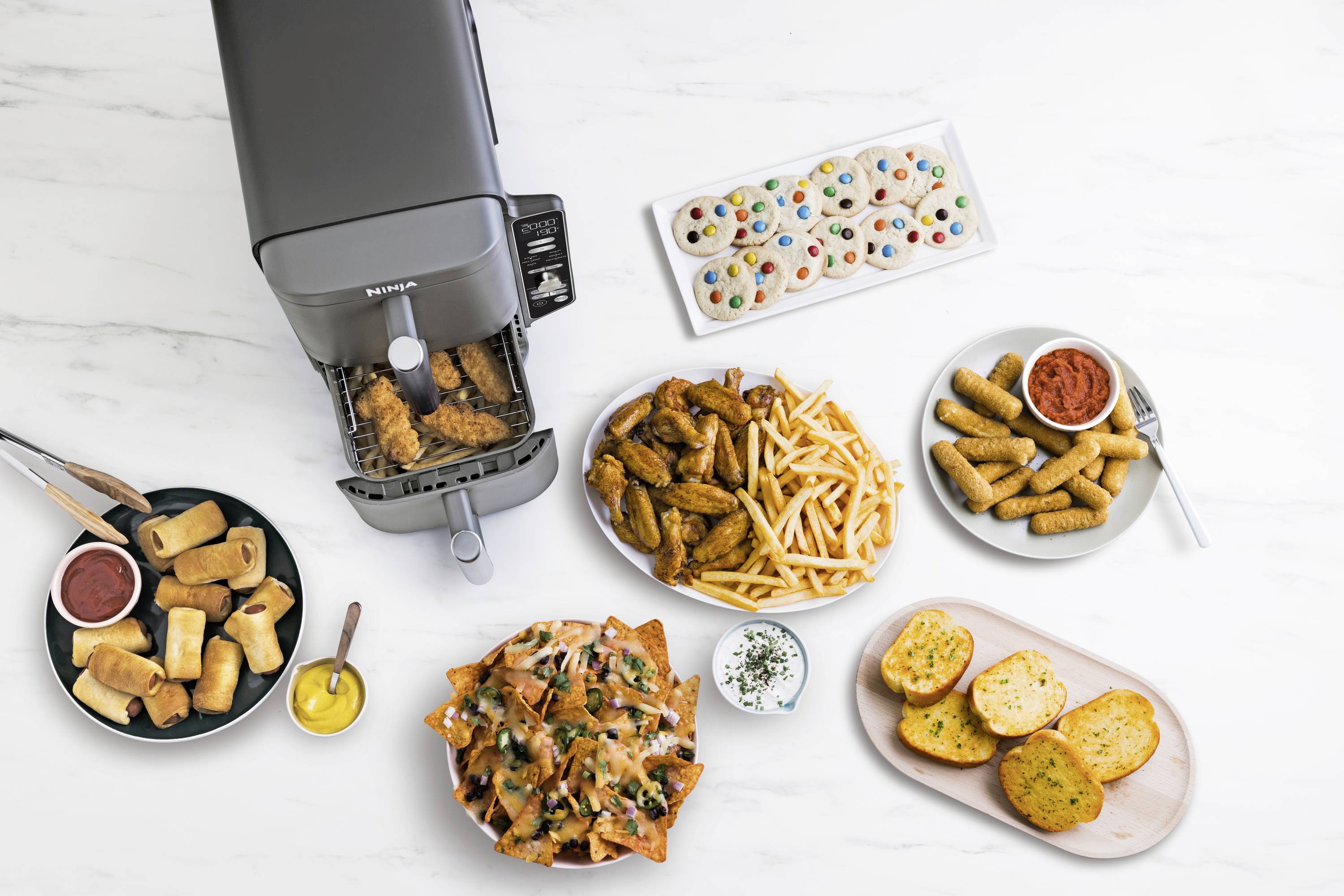 Různé snacky na stole: kuřecí křídla, hranolky, jarní závitky, sýrové nachos, sušenky a chléb poblíž horkovzdušné fritézy.