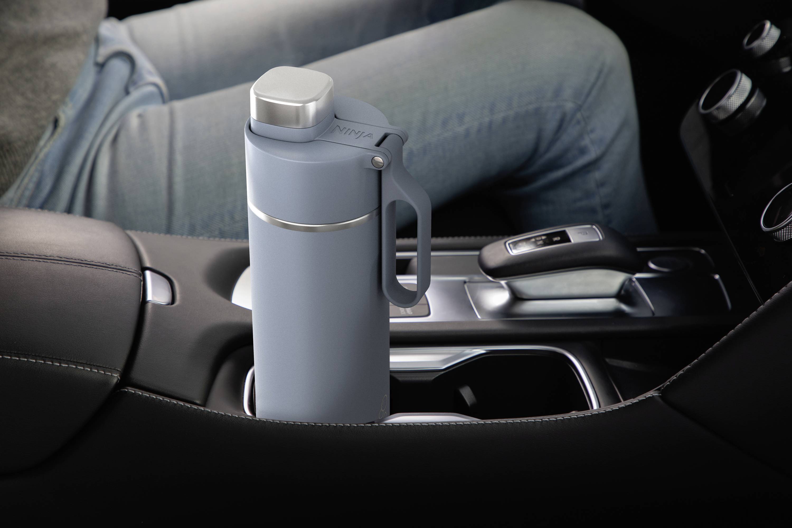 Šedá termoska stojí v držáku nápojů automobilu vedle řadicí páky. Osoba sedí na sedadle řidiče.