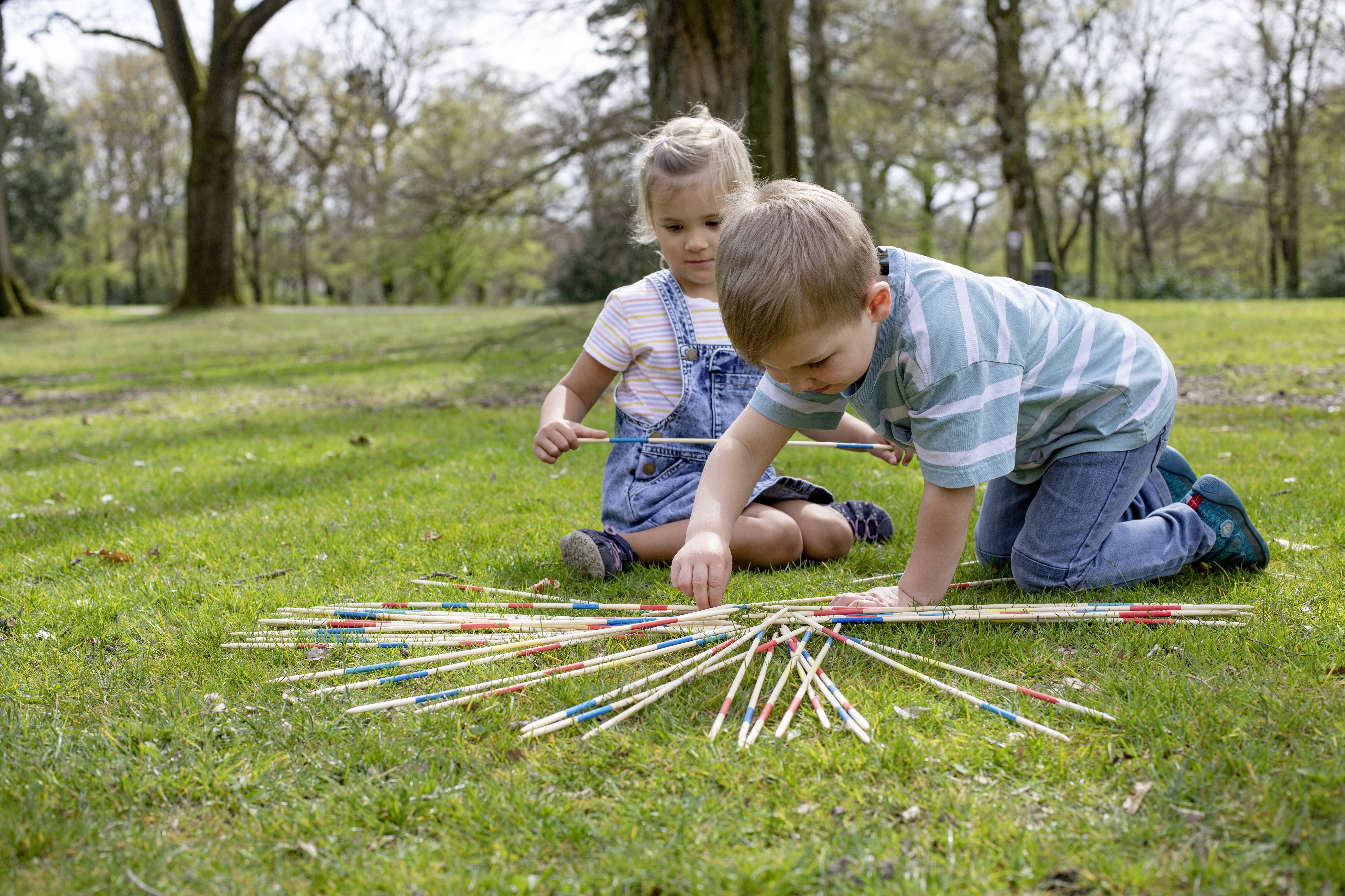 Dvě děti hrají hru Pick-up Sticks v parku. Klečí na trávě, obklopeny stromy. Soustředěné výrazy ve tváři při hře.