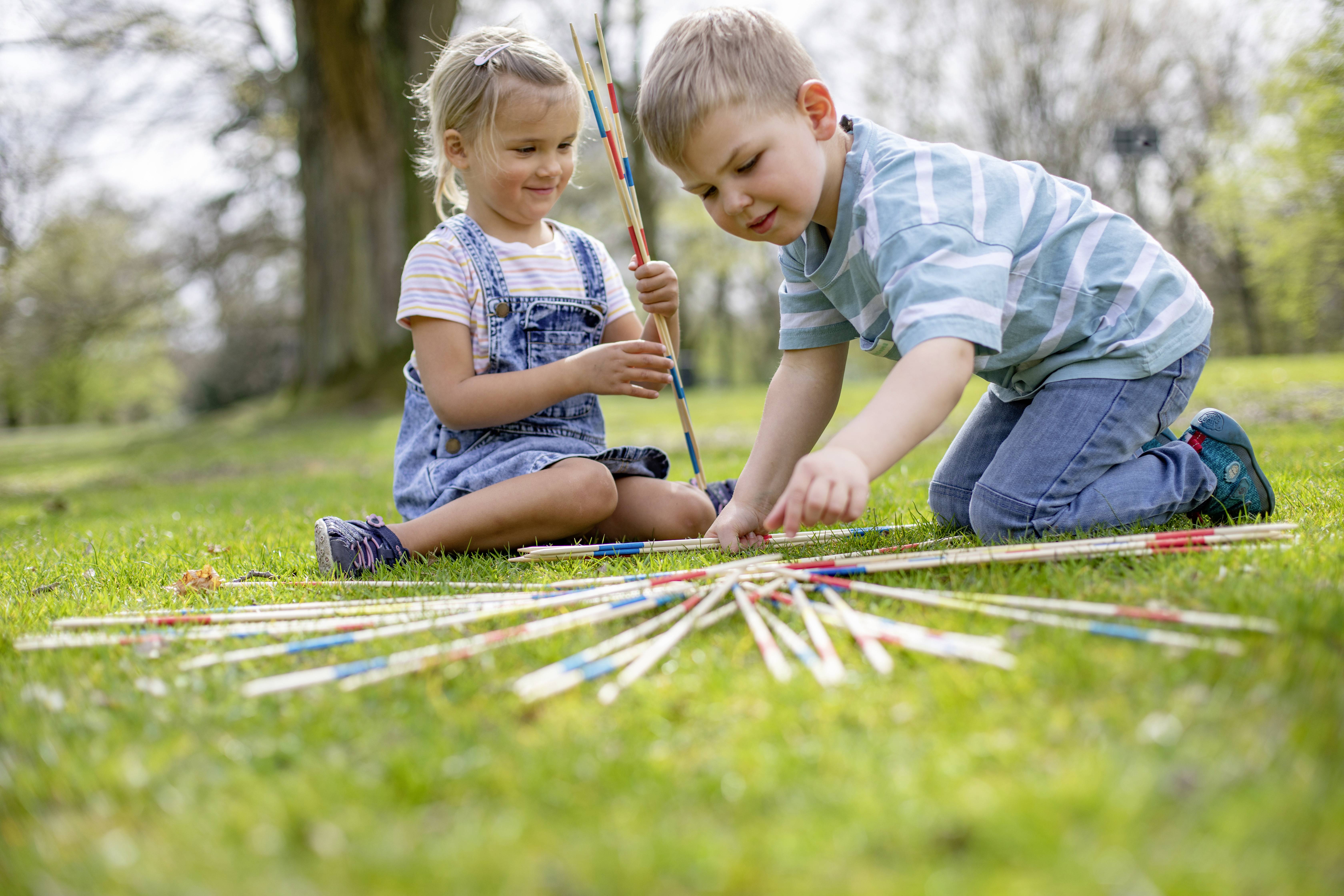 Dvě děti si hrají na louce s pick-up-sticks. Dívka sbírá tyčky, zatímco chlapec jednu zvedá. Je slunečný den.