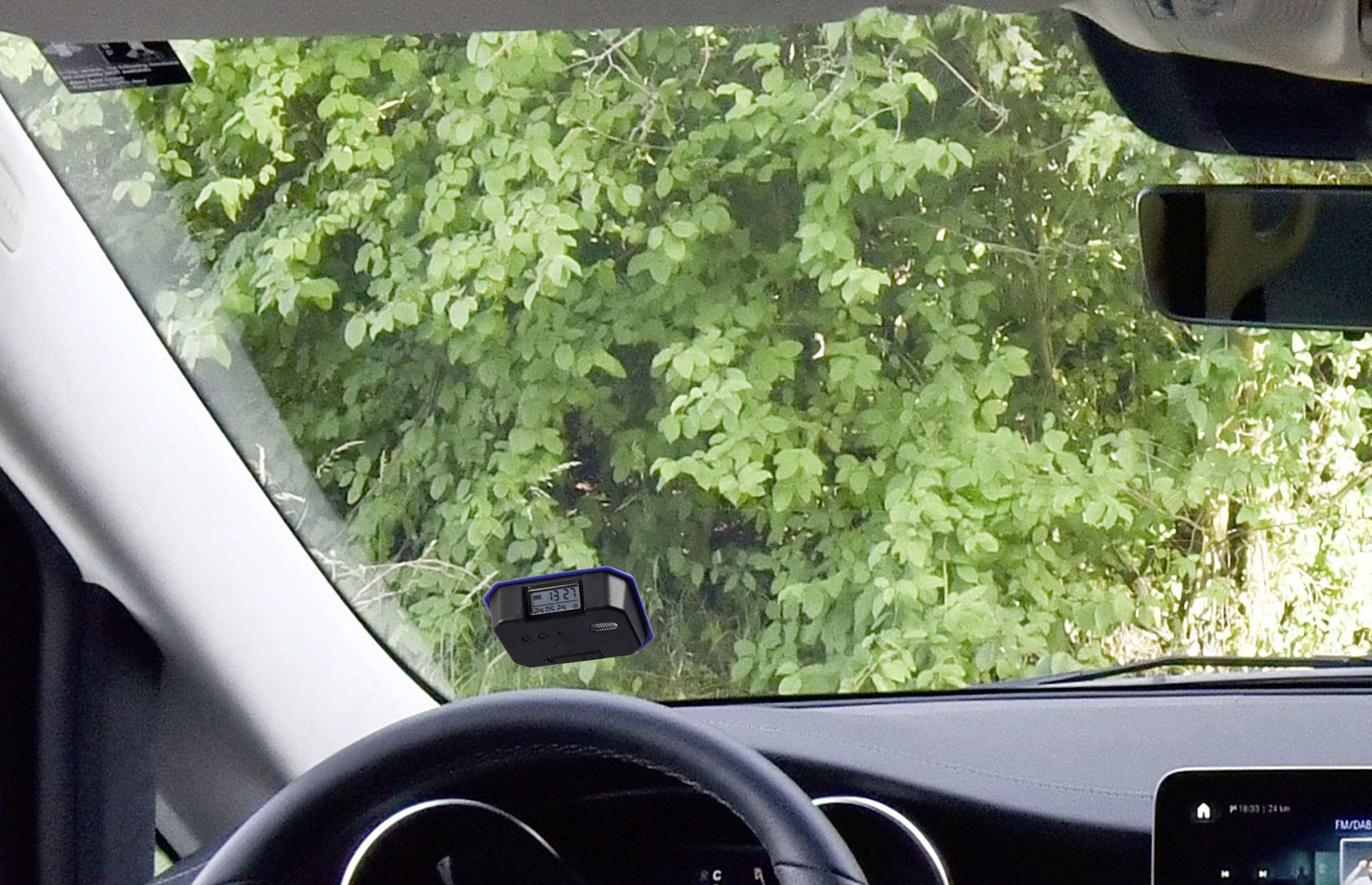 Pohled z auta skrz čelní sklo na zelené keře. Viditelný volant a digitální zařízení.