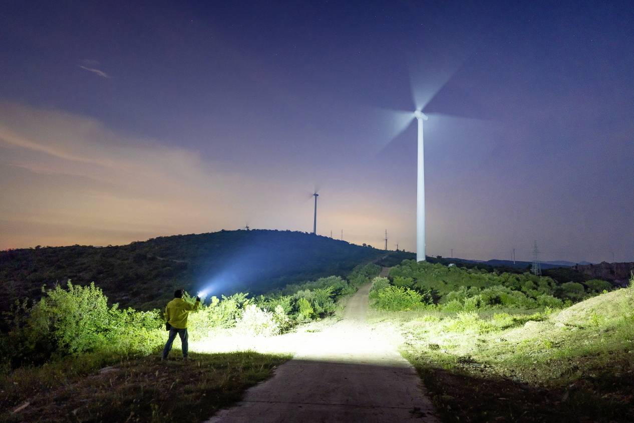 Osoba s baterkou stojí za soumraku před osvětlenou větrnou turbínou na kopci, obklopená dalšími větrnými mlýny.