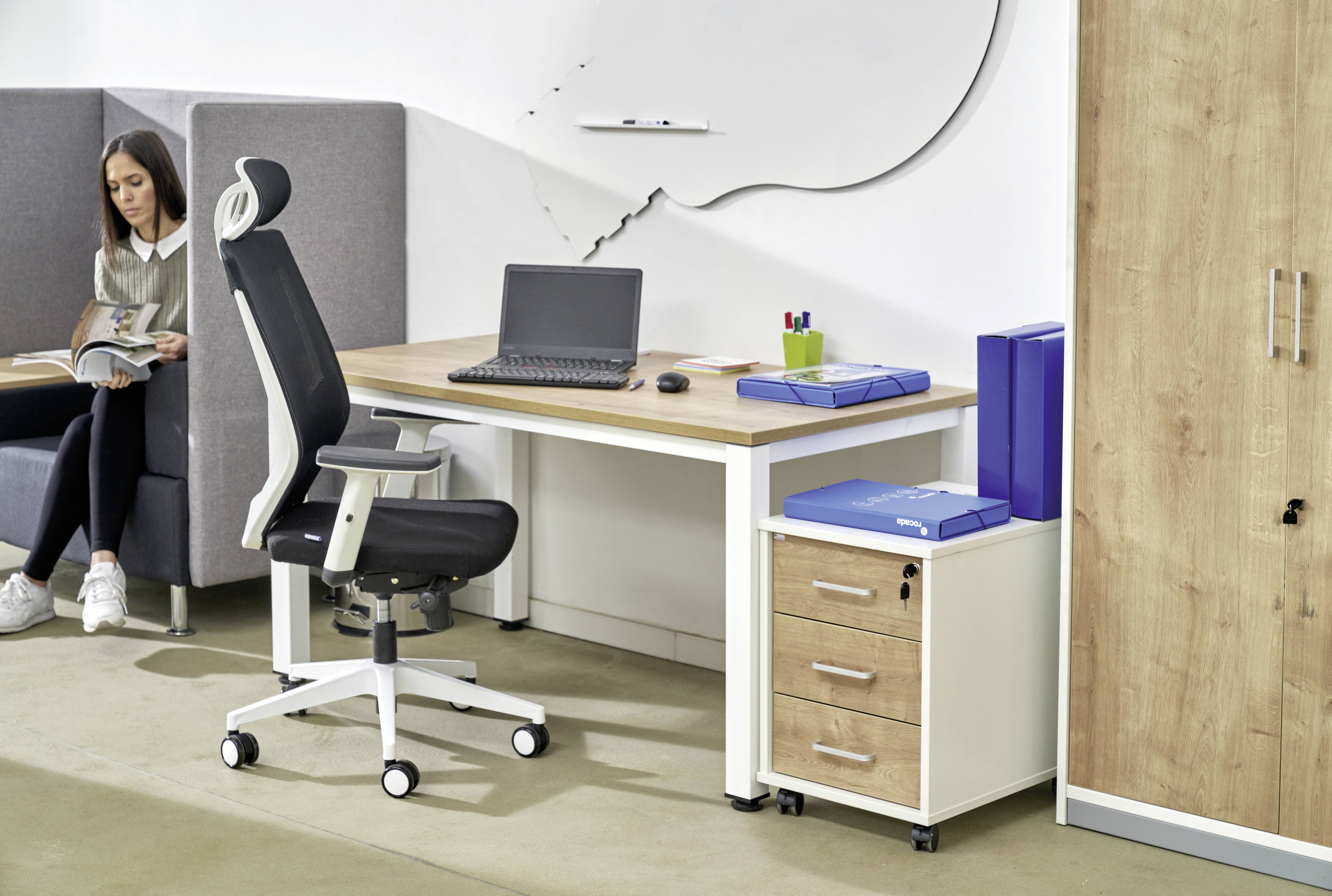 Moderní kancelář s psacím stolem, na kterém je položen laptop. Vedle něj stojí ergonomická kancelářská židle a mobilní kontejner. V pozadí sedí žena na pohovce a čte knihu.