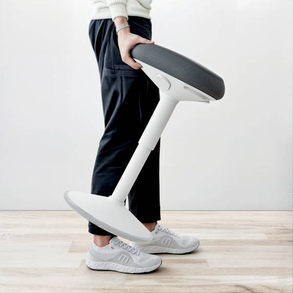 Osoba drží moderní ergonomickou židli se šikmým sedákem v světlém prostoru. Židle má minimalistický design.