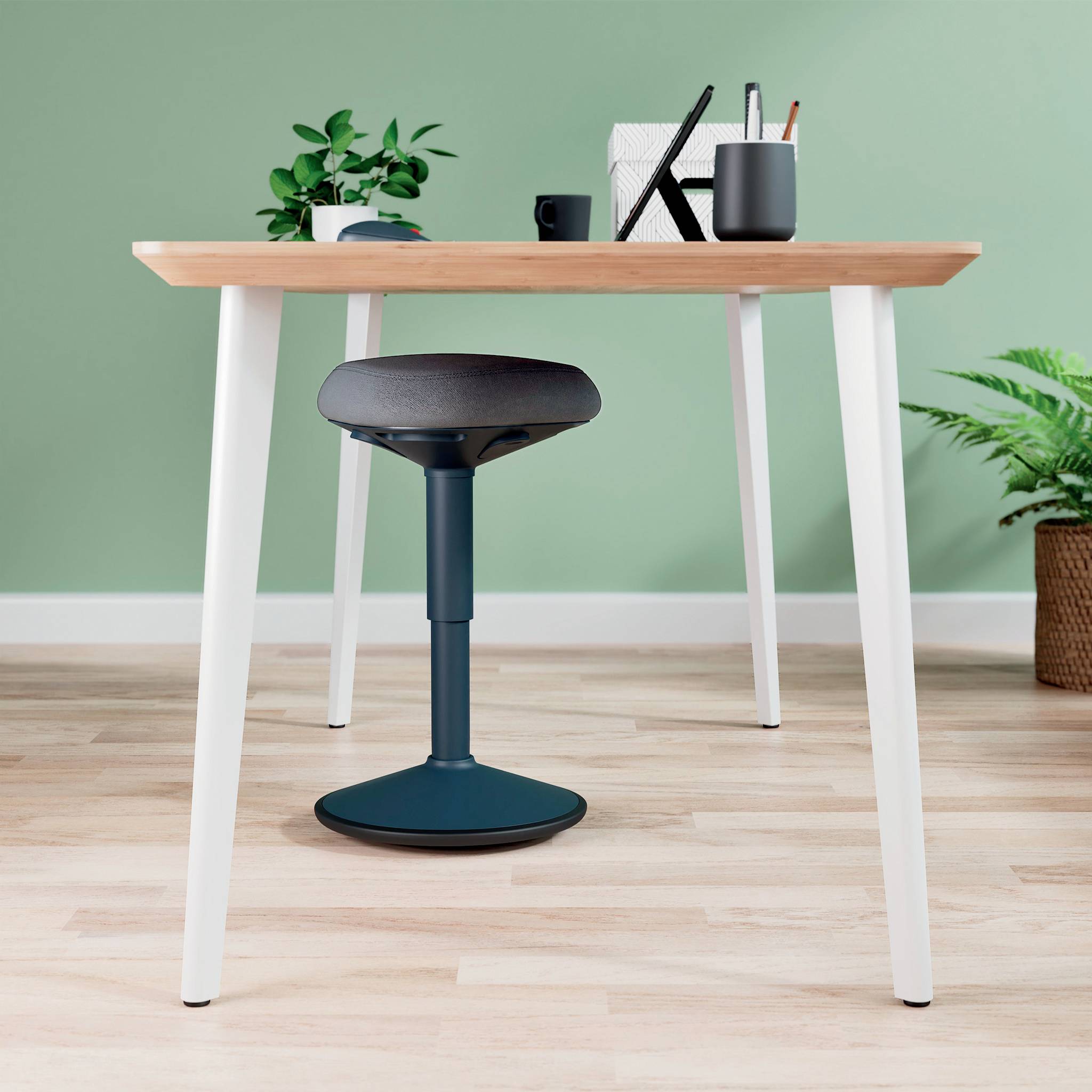 Moderní psací stůl s zeleným pozadím, na němž leží psací potřeby, poznámky a malý květináč. Pod stolem stojí ergonomická stolička.