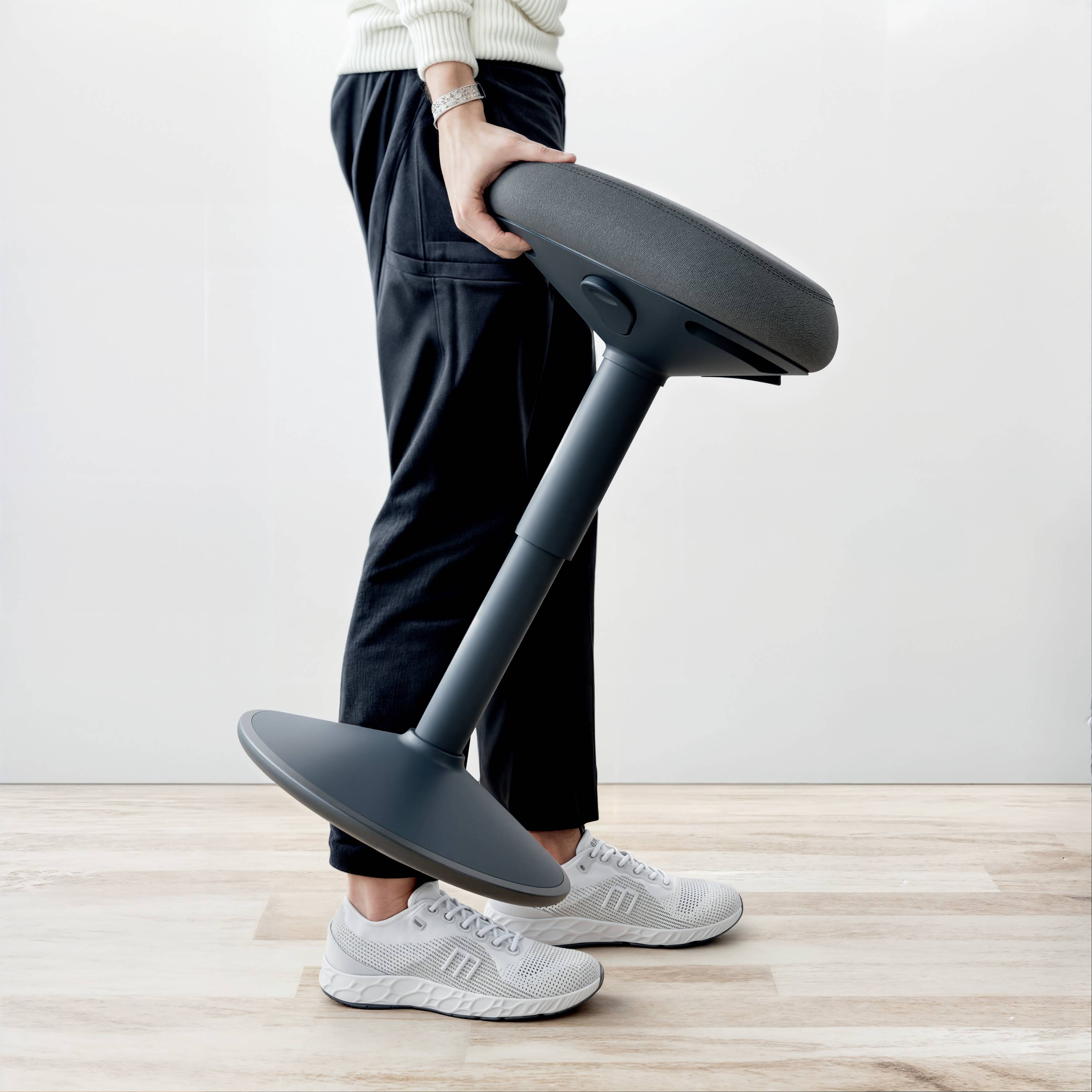Osoba drží moderní, nastavitelnou židli se šikmým sedákem v světlém prostoru. Židle má kulatou základnu a umožňuje ergonomické sezení.