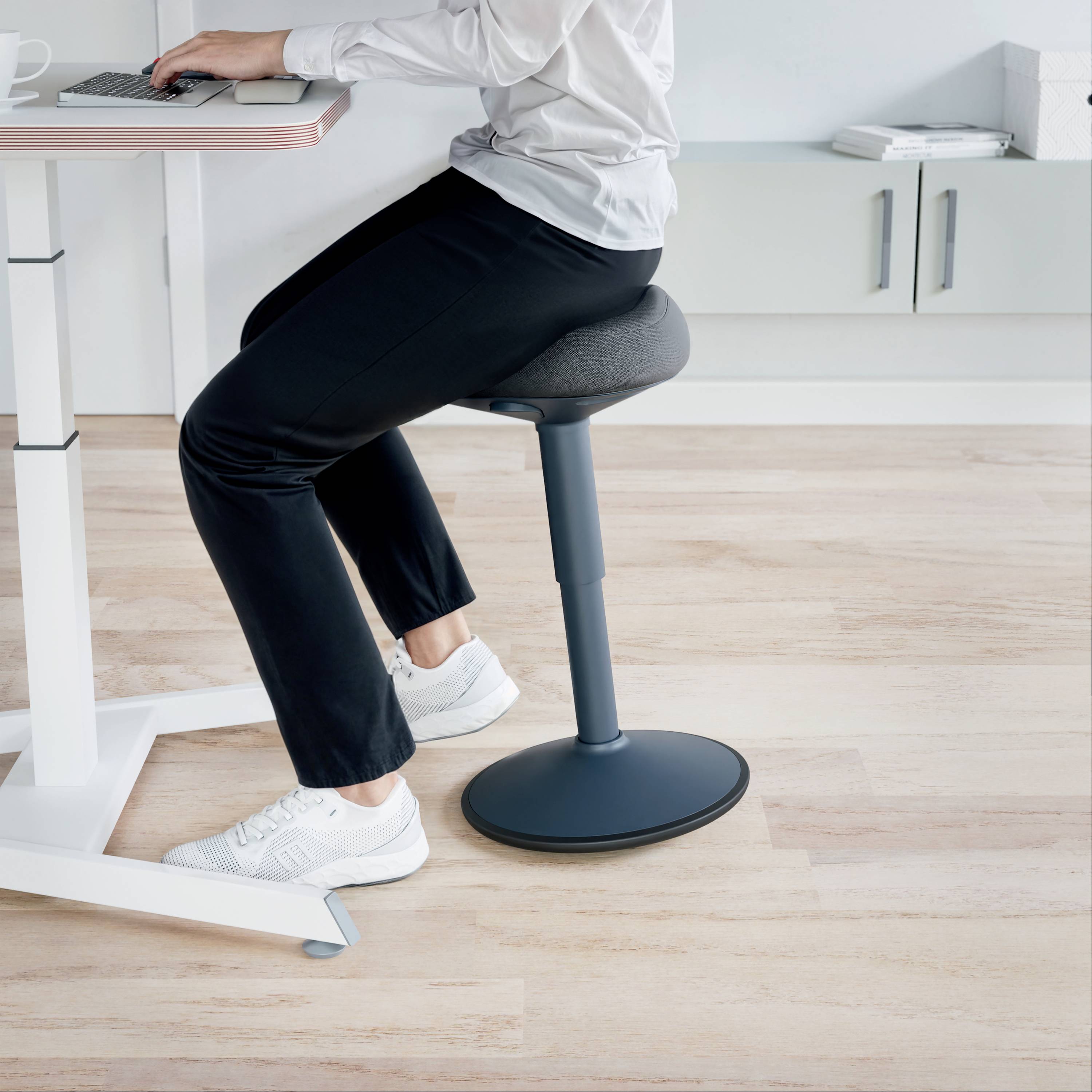 Osoba sedí na ergonomické stoličce u výškově nastavitelného psacího stolu, ruce na klávesnici, v moderním kancelářském prostředí.