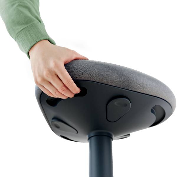 Osoba v zeleném svetru drží šedou stoličku s nastavitelným sedákem, která nabízí ergonomické funkce.