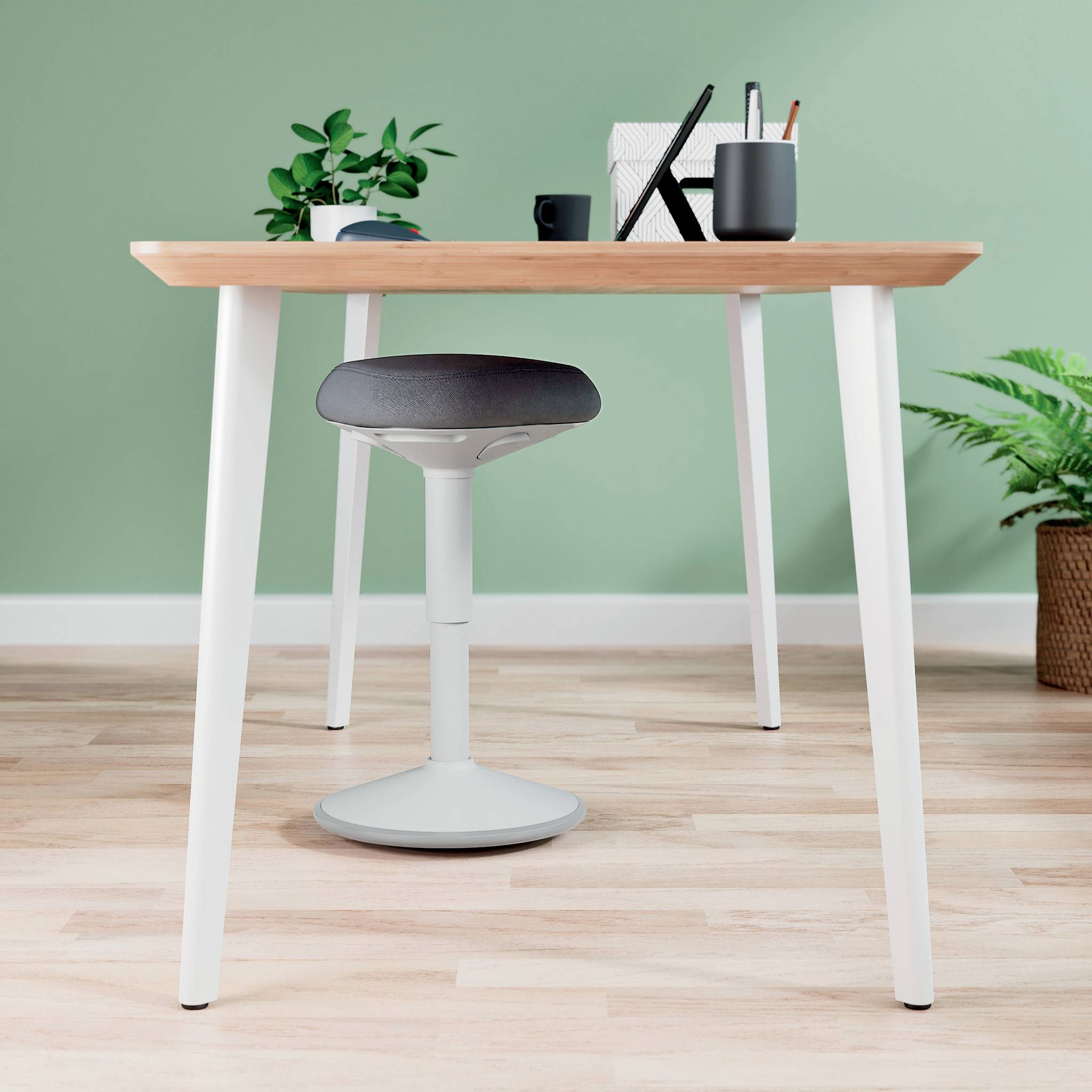 Psací stůl s dřevěnou deskou, kancelářské rostliny, kancelářské potřeby a ergonomická stolička na světlé dřevěné podlaze před zelenou stěnou.