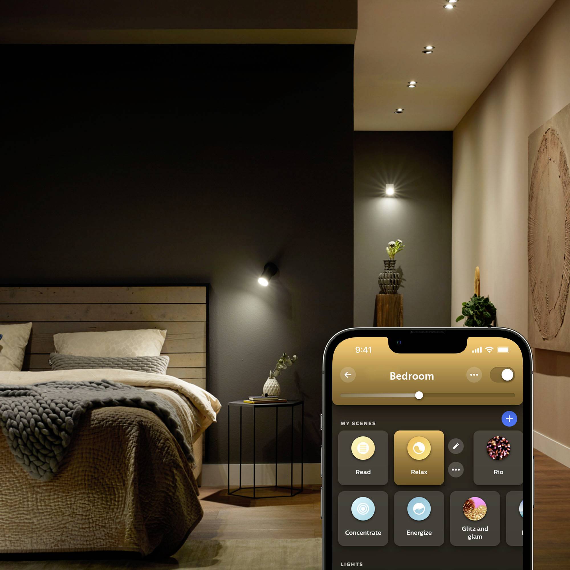 Smartphone zobrazuje aplikaci s ovládacími možnostmi osvětlení ložnice: 'Čtení', 'Relaxace', 'Romantika'. V pozadí světlá, moderně zařízená ložnice.