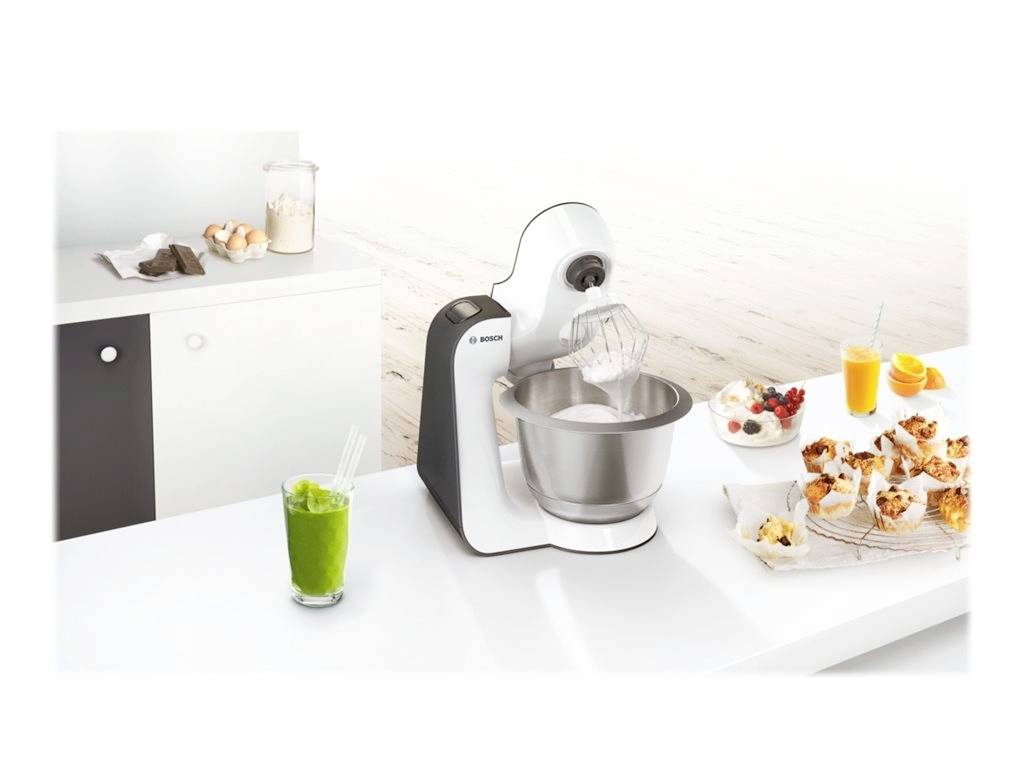 Kuchyňský robot na stole při hnětení těsta. Vedle sklenice se zeleným smoothie, muffiny, pomerančový džus a další nápoj.